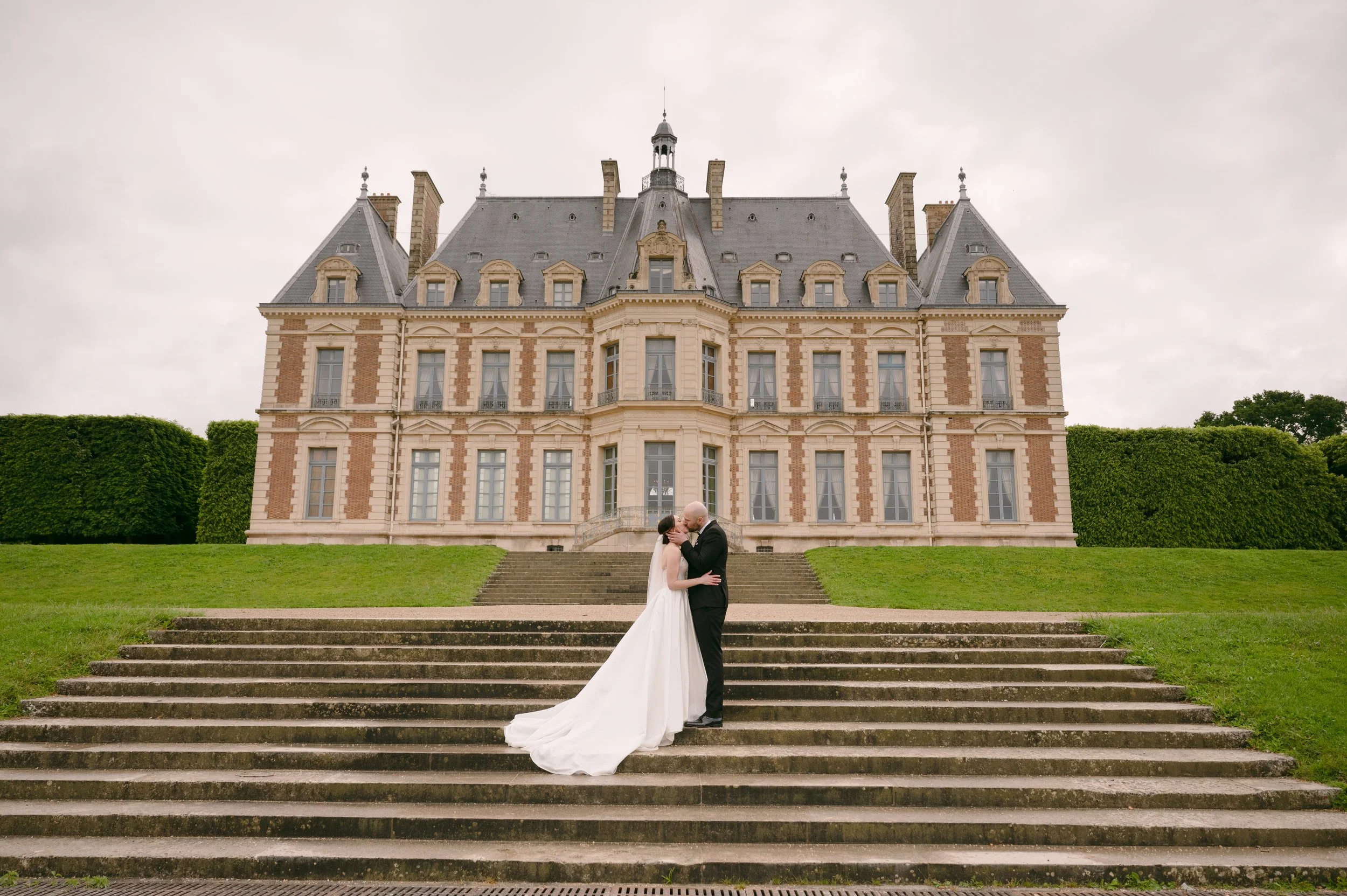 A bride and groom kiss on the steps of a historic chateau or castle, surrounded by green grass and bushes, on a cloudy day.