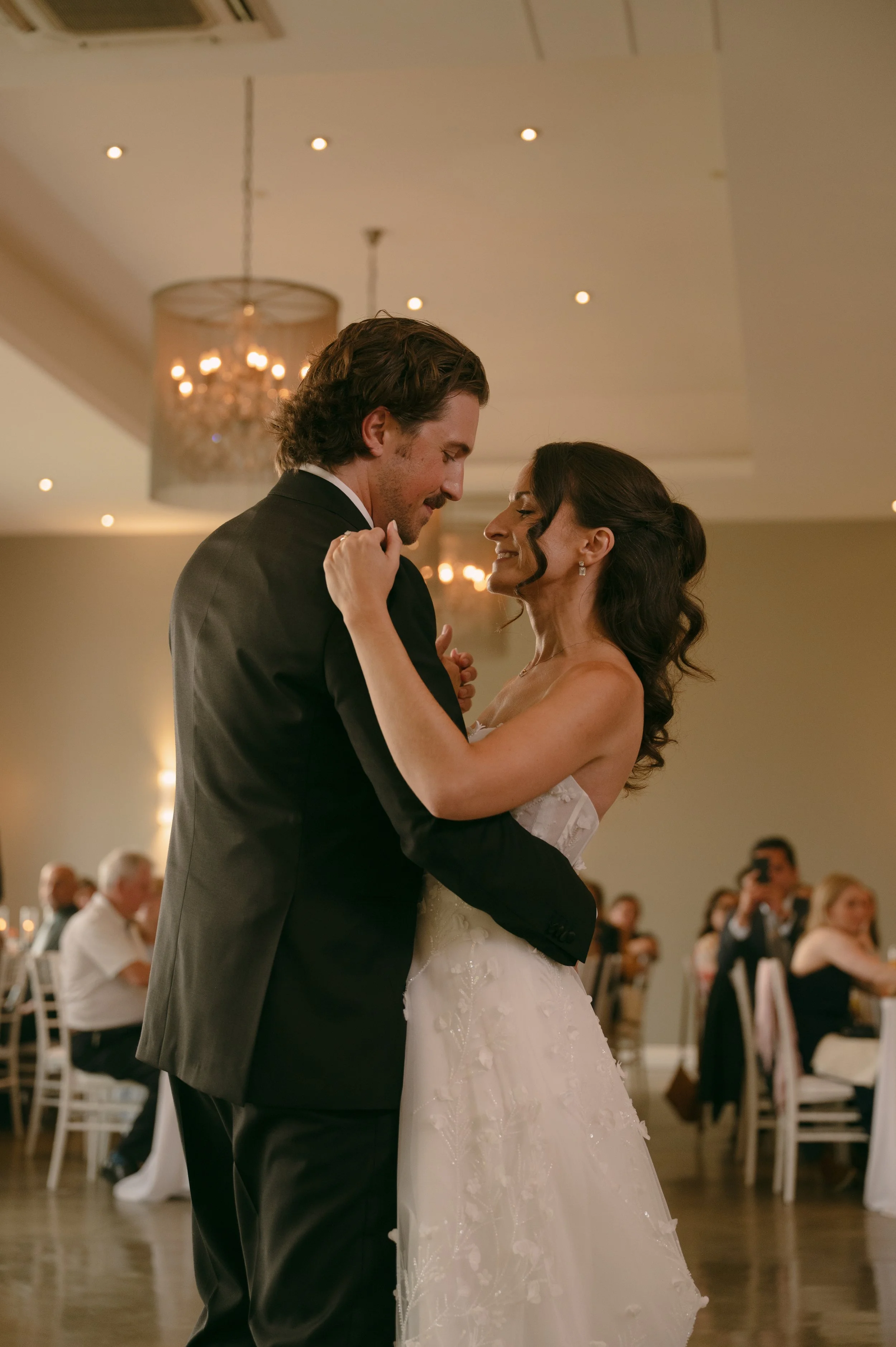 A bride and groom share a dance at their wedding reception, smiling and looking into each other's eyes. Guests are seated in the background.