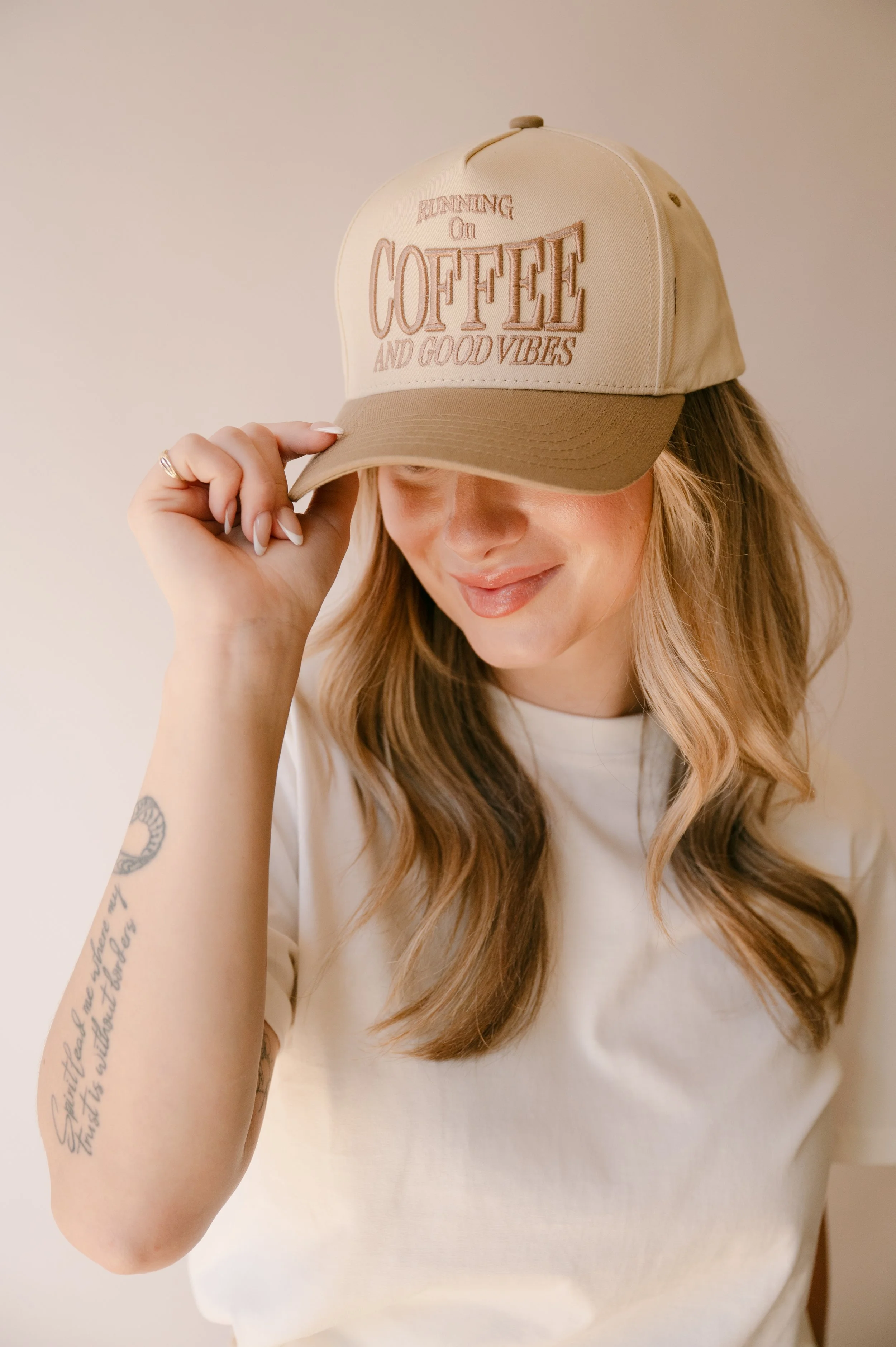 A woman wearing a beige baseball cap with embroidered text that reads 'Running on Coffee and Good Vibes' and a white t-shirt, smiling softly, with long wavy hair and visible tattoos on her left arm.