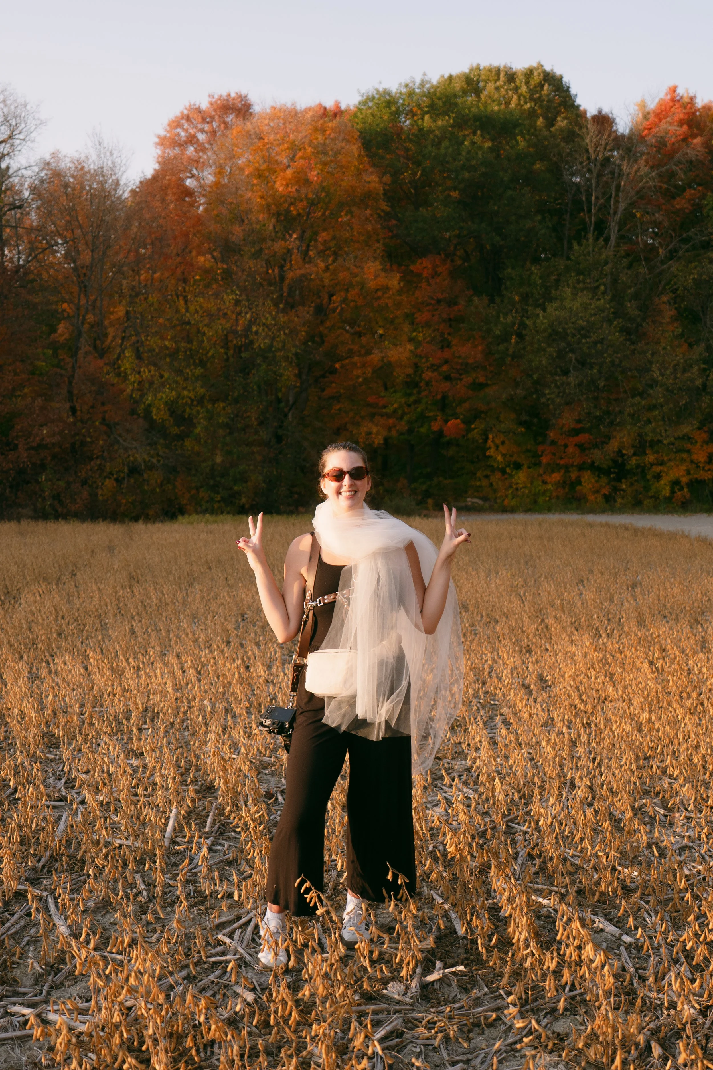A woman in black pants, sunglasses, and a white scarf stands in a dry, brown field with tall trees displaying fall foliage in the background, smiling and flashing peace signs.