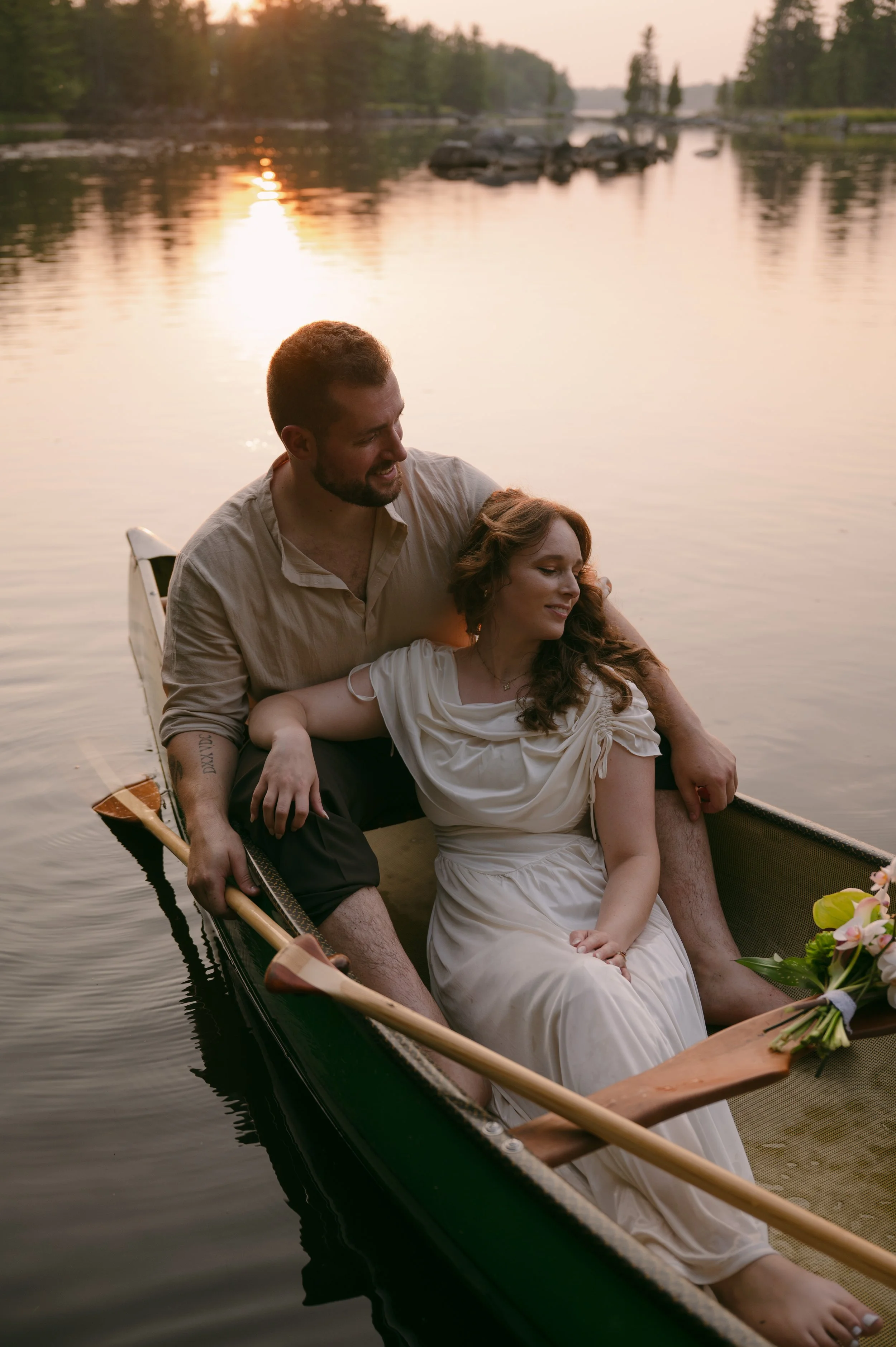 A couple sitting in a canoe on a lake at sunset, with the man smiling and the woman relaxing with her eyes closed, surrounded by a calm water and trees in the background.