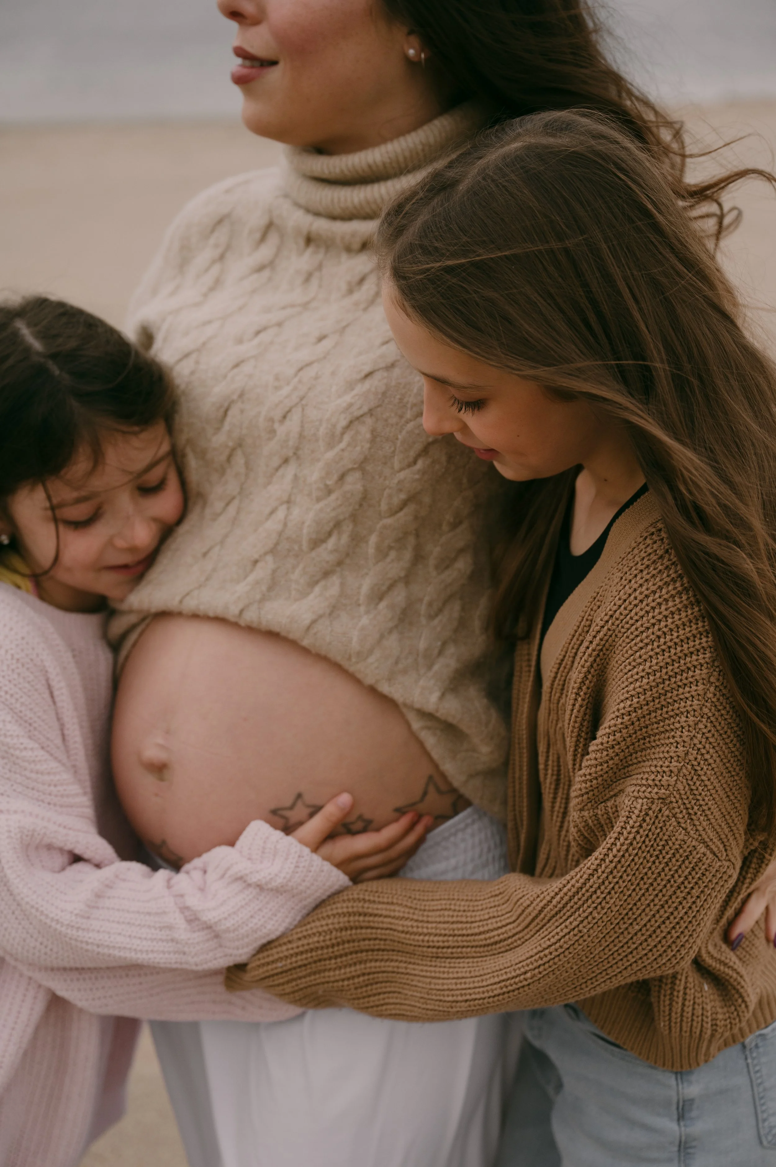 A pregnant woman with two young girls hugging her belly on a beach.