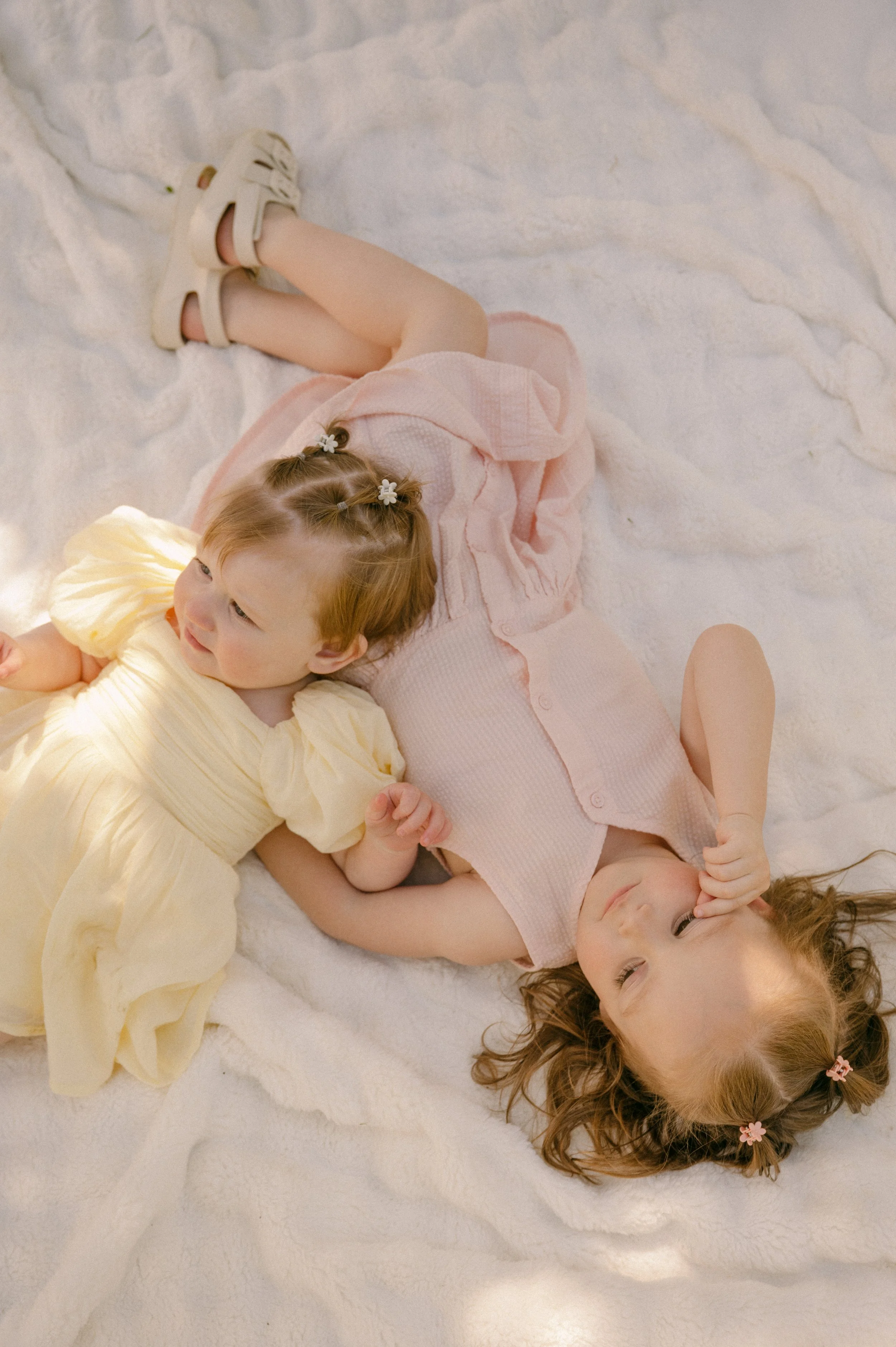 Four young children lie on a soft white blanket, smiling and looking relaxed. They are dressed in light pastel-colored clothes, with two girls having hair styled with small hair clips and flowers. The mood is joyful and peaceful.