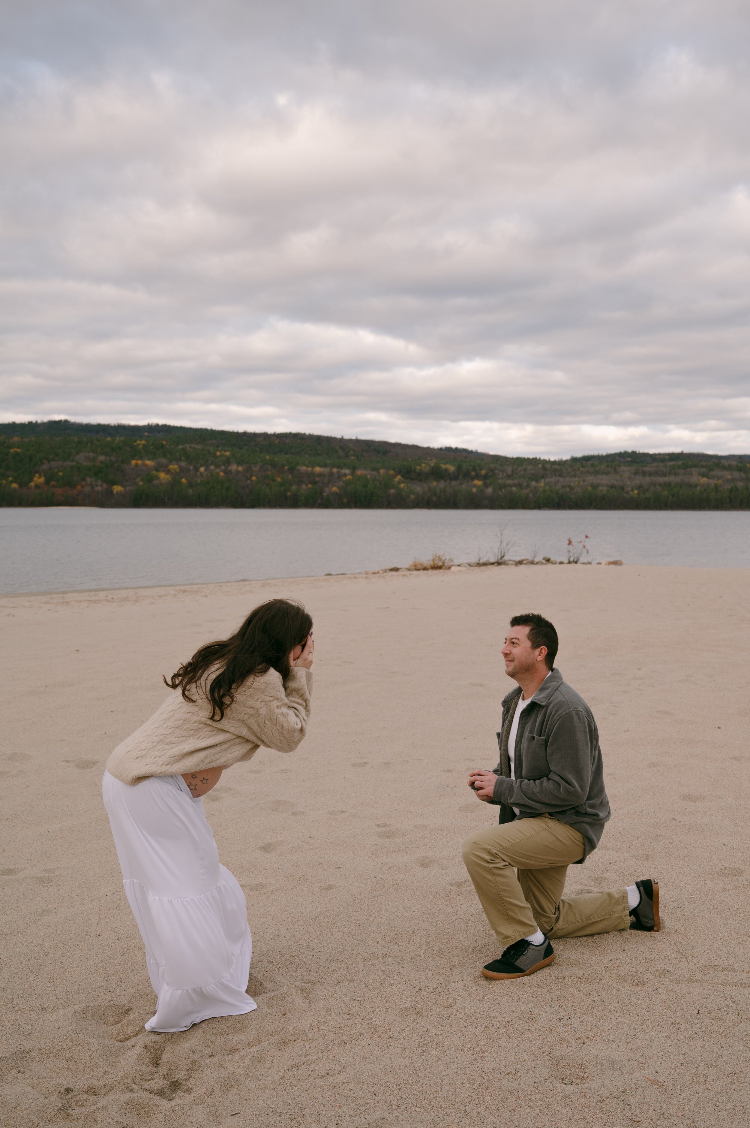 A man proposing marriage to a woman on a sandy beach with water and hills in the background on a cloudy day.