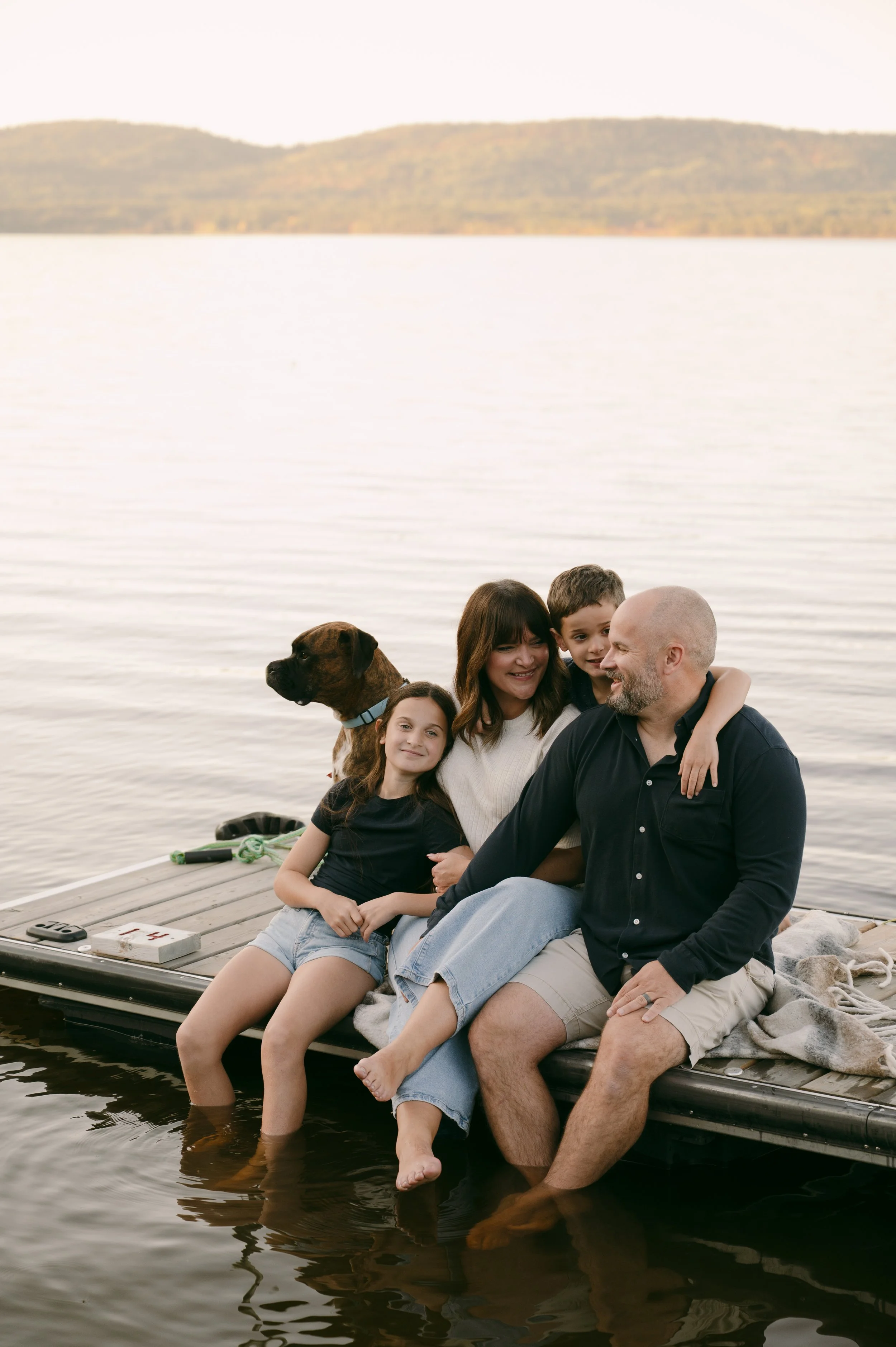 Family sitting on a dock by a lake, smiling, with a dog, during sunset or early evening.
