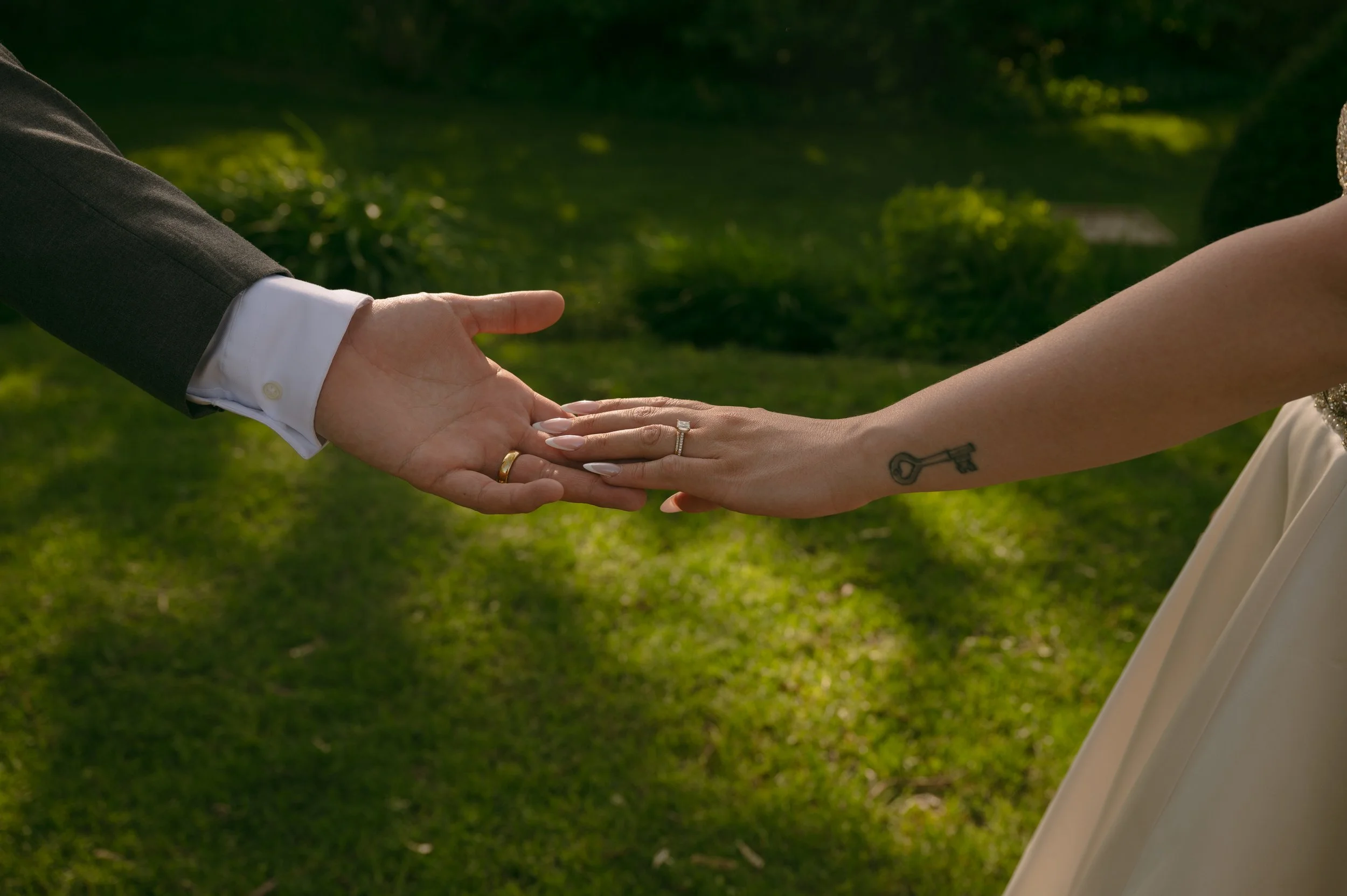 A close-up of a bride and groom's hands holding each other, with a wedding ring on each hand, outdoors with greenery in the background.
