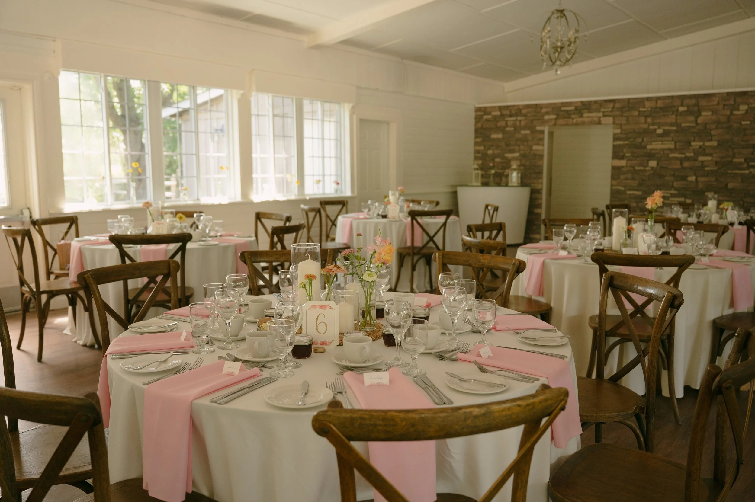 A elegantly decorated banquet hall with round tables covered in white tablecloths and pink table runners, set with glassware, plates, and silverware, and adorned with small floral centerpieces, ready for a celebration.