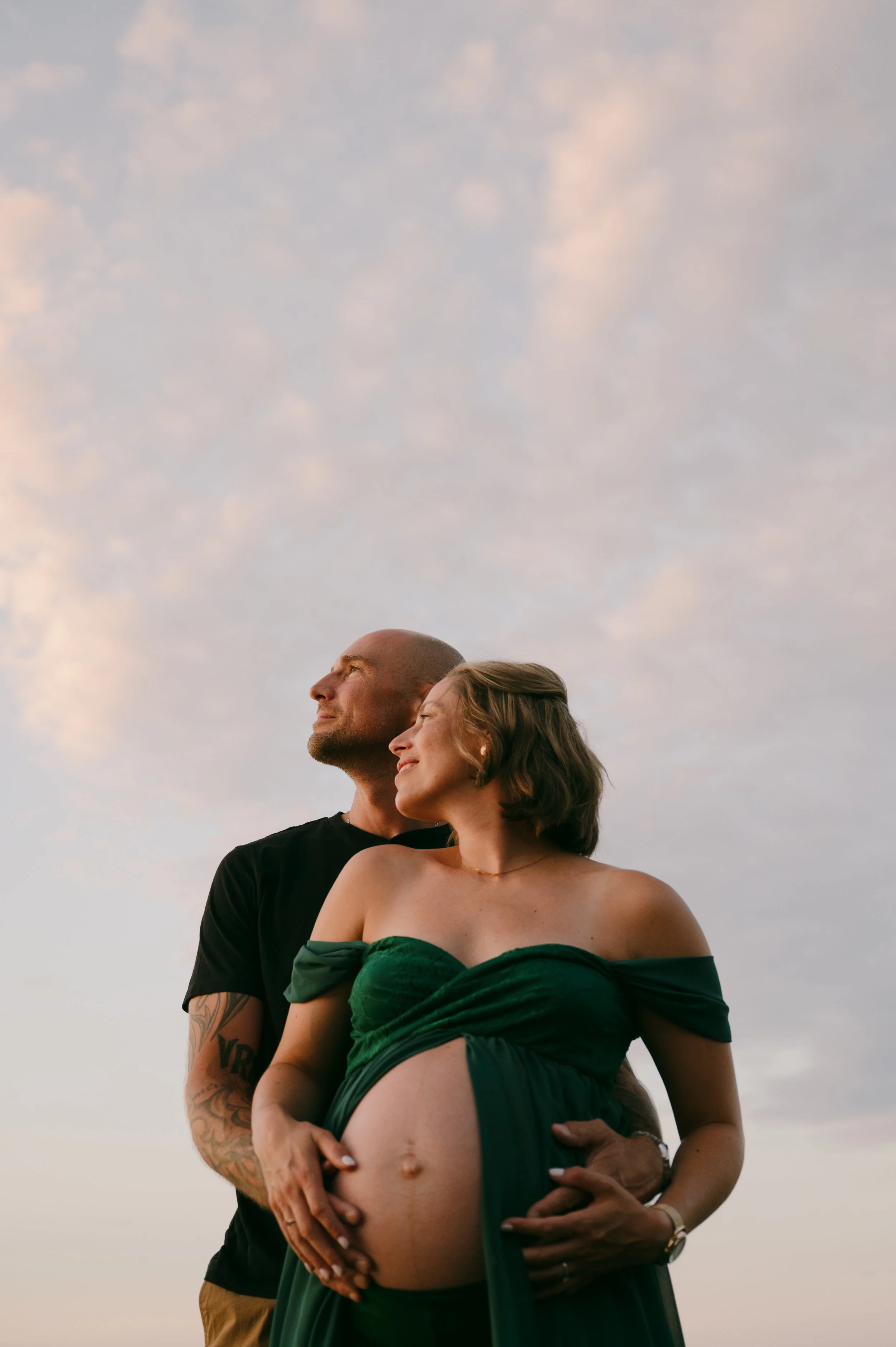 A pregnant woman in a green off-shoulder dress holding her belly, standing in front of a man with a tattooed arm, both looking content against a sky with soft clouds during sunset.