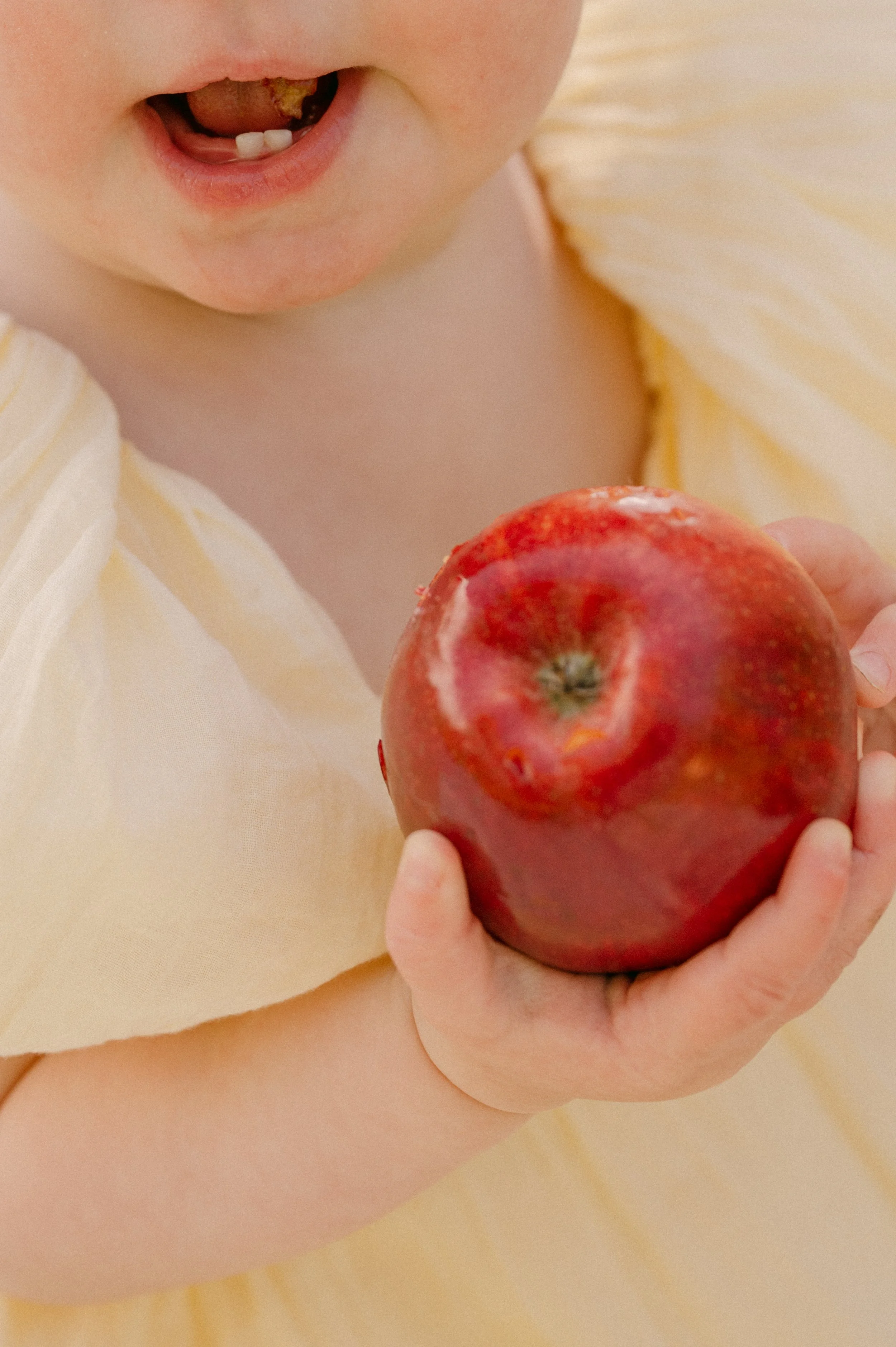 Close-up of a young child holding a red apple, with part of their face and neck visible, wearing a light yellow and white striped shirt.
