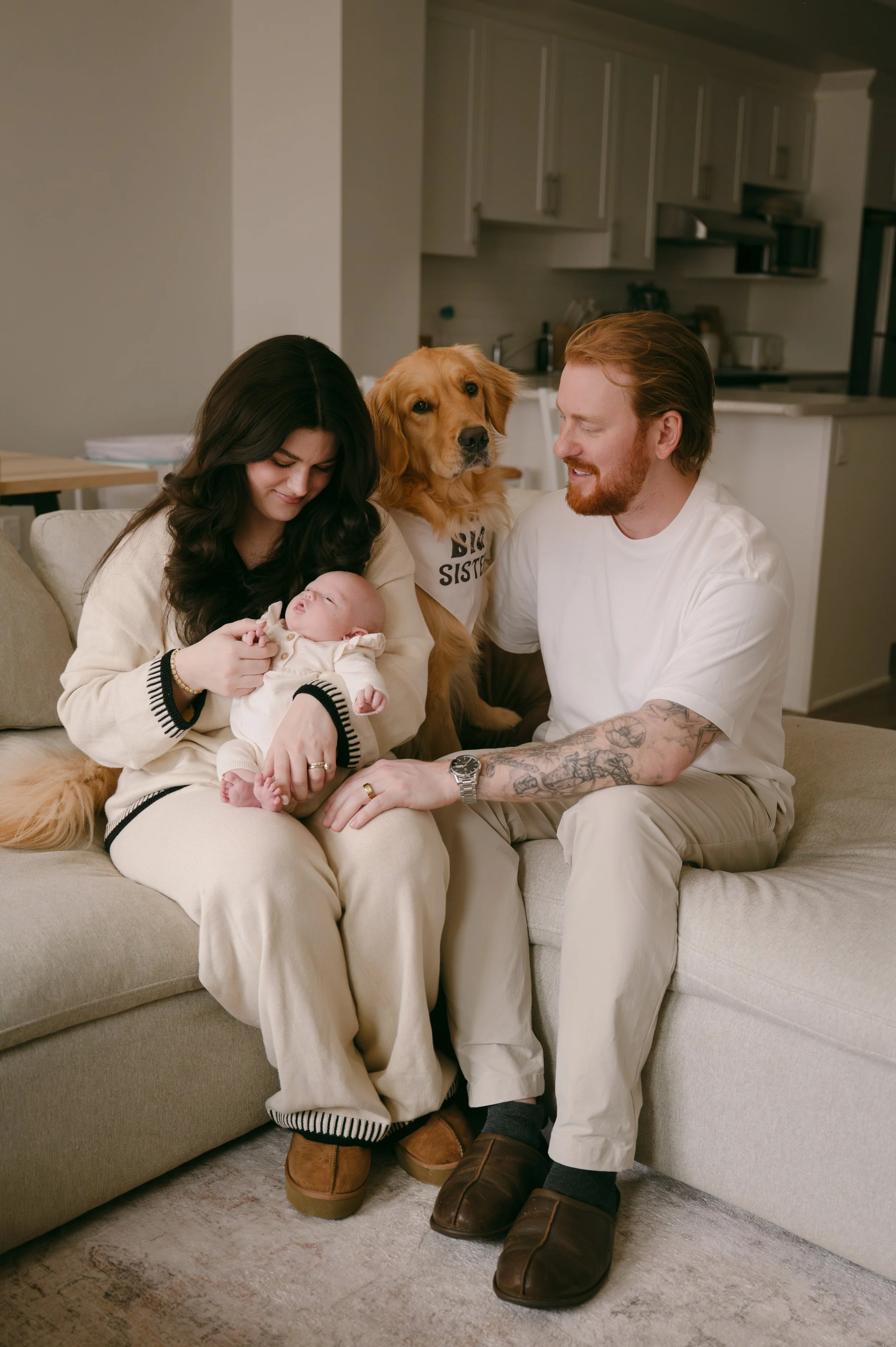A family sitting on a beige couch in a living room, consisting of a woman holding a baby, a man with tattoos, a golden retriever dog sitting between them, and a background of kitchen cabinets.
