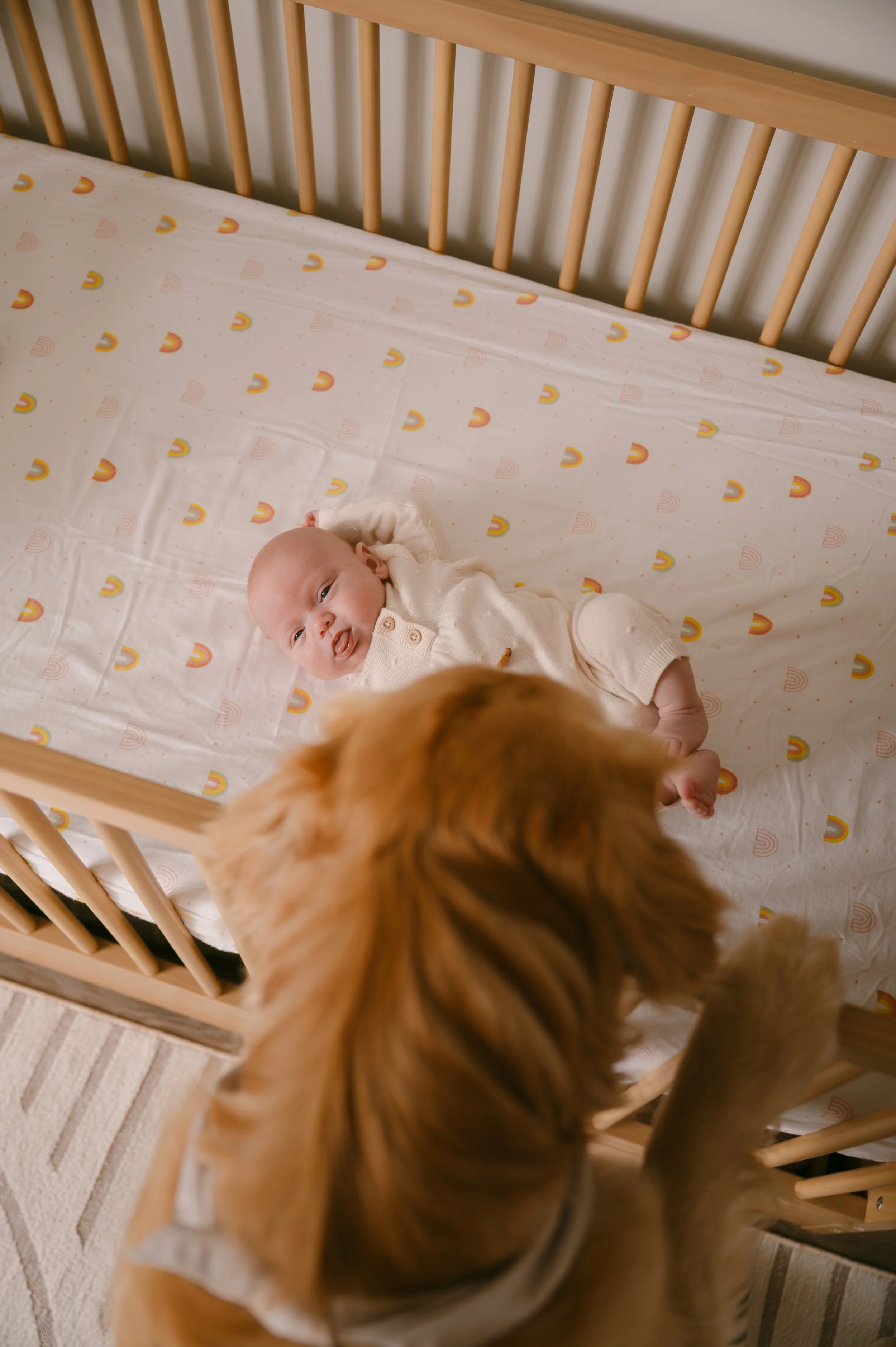 A baby lying in a crib with a patterned sheet, looking up at a dog whose head is visible at the bottom of the image.