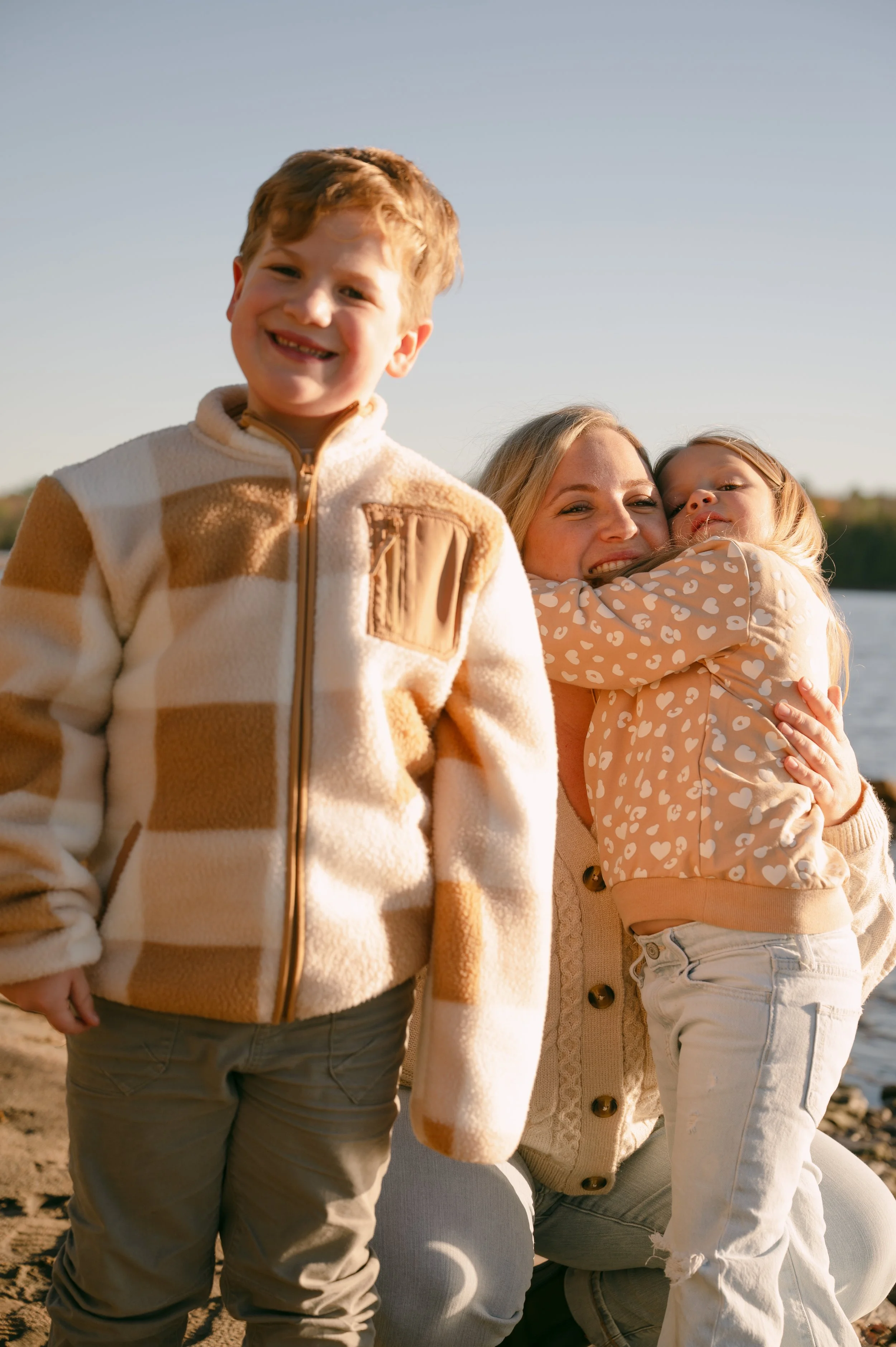 A woman with blonde hair sitting on the ground hugging a young girl with blonde hair and wearing a heart-patterned jacket, while a young boy with red hair and a checkered fleece jacket stands nearby smiling.