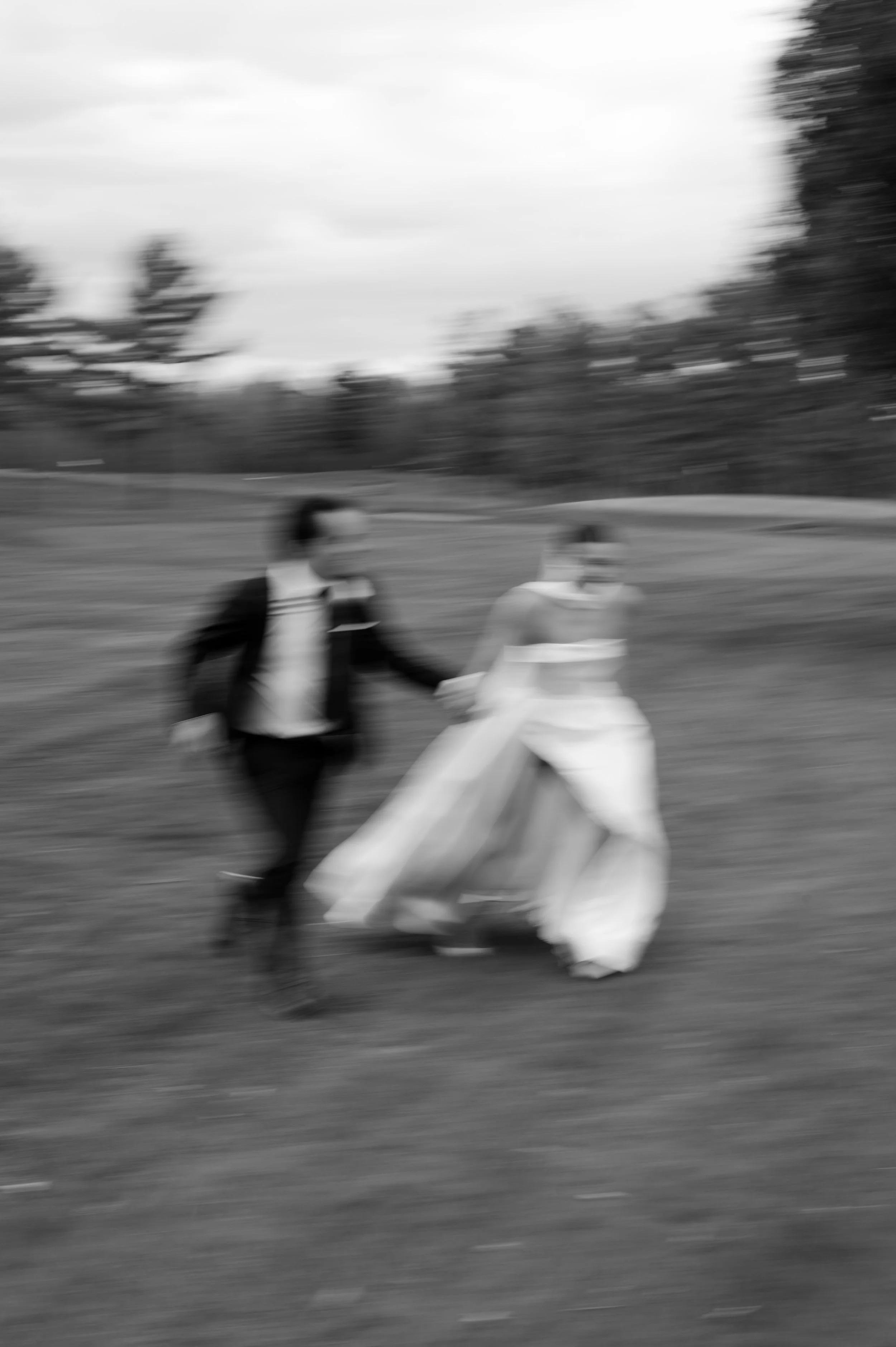 A blurry black and white photo of a young couple walking outdoors in a park or field, with the woman wearing a wedding dress and the man in a dark suit, holding hands.