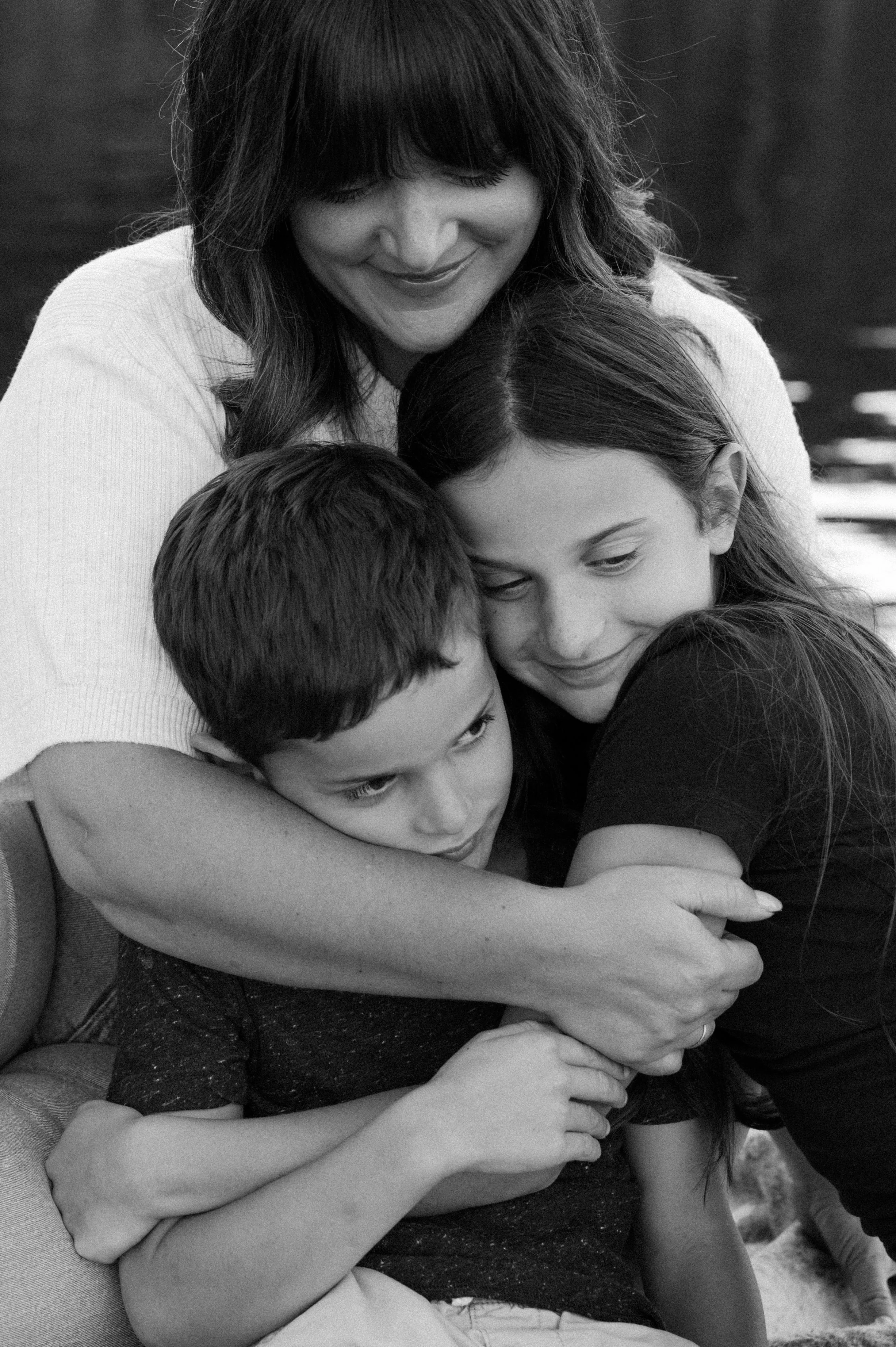 A woman hugging a young girl and a young boy, all smiling and pressing their faces together in a warm embrace.