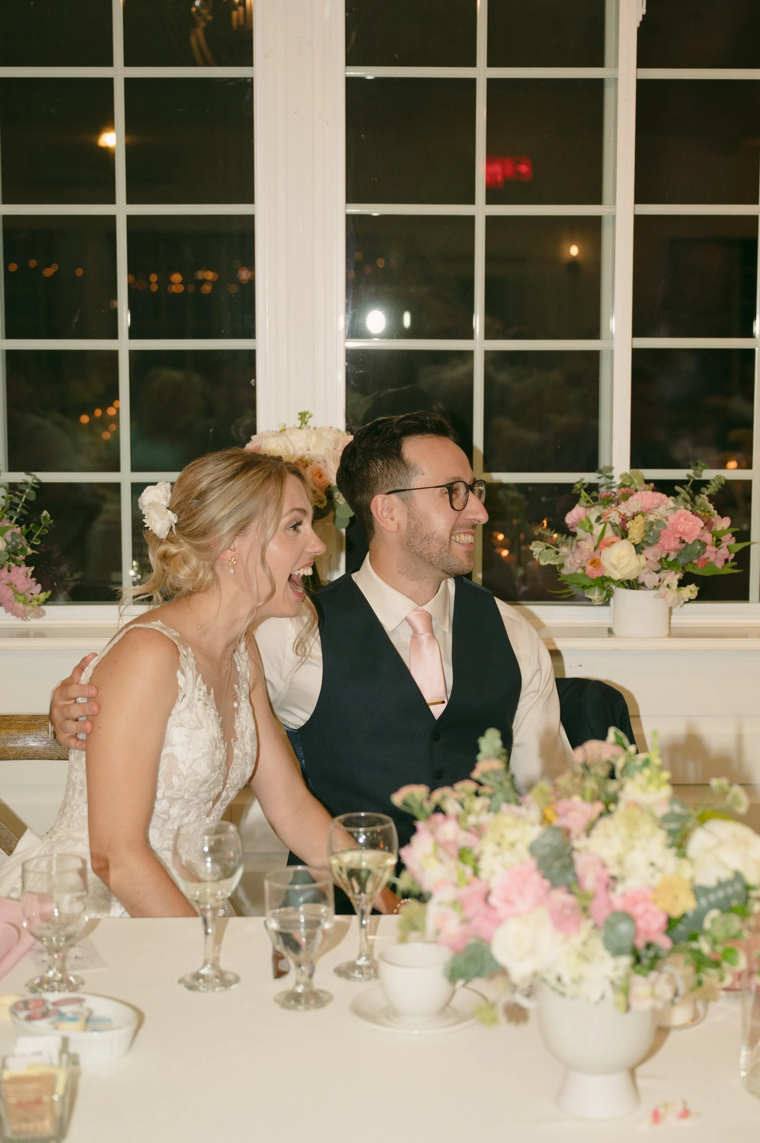A bride and groom sitting at a wedding reception table, smiling and laughing. The bride is wearing a lace wedding dress and has her hair styled in loose waves with a flower in her hair. The groom is wearing glasses, a white shirt, a dark vest, and a 