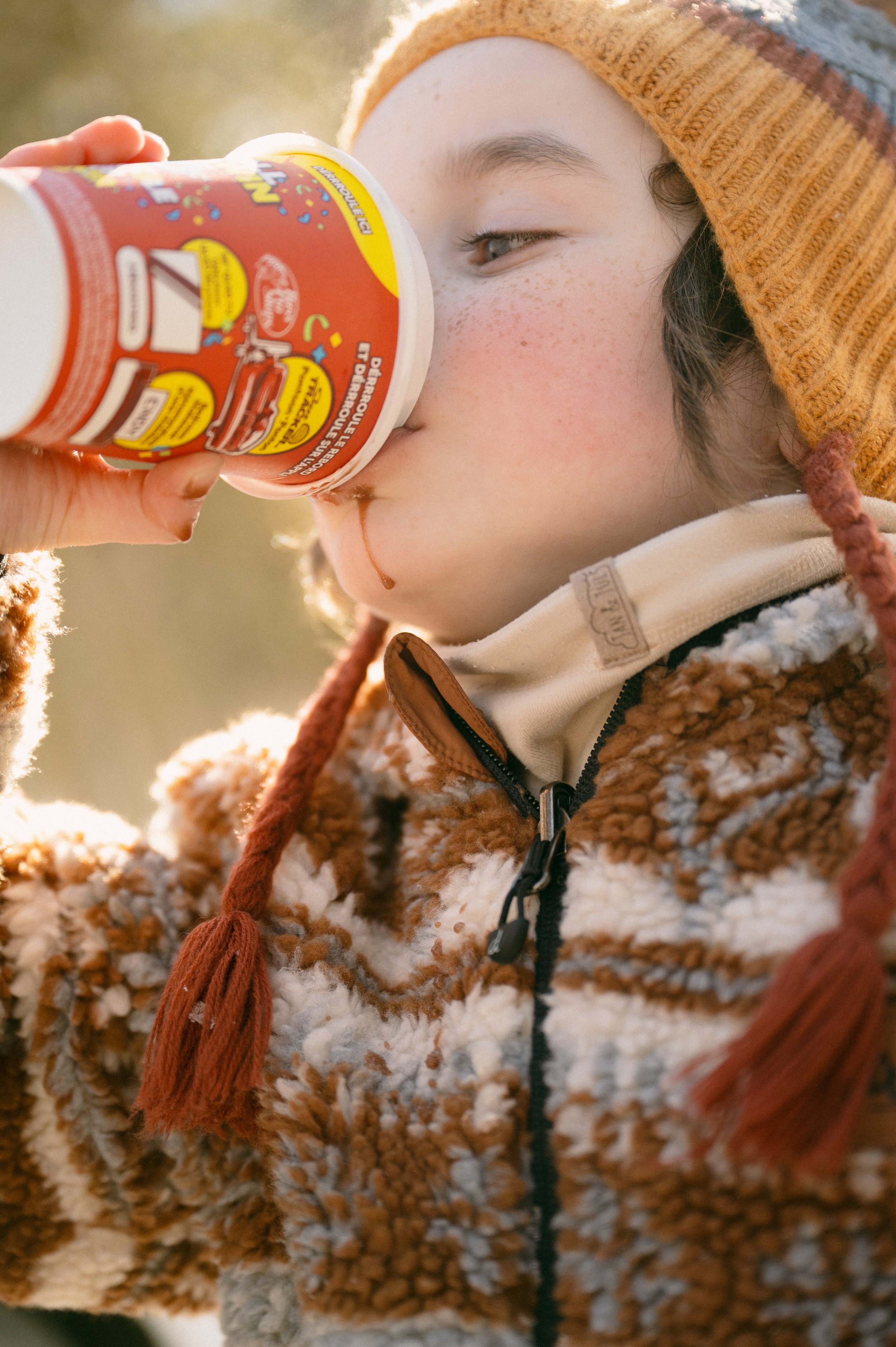 A young person wearing a knit hat and fleece jacket drinking from a colorful can outdoors.