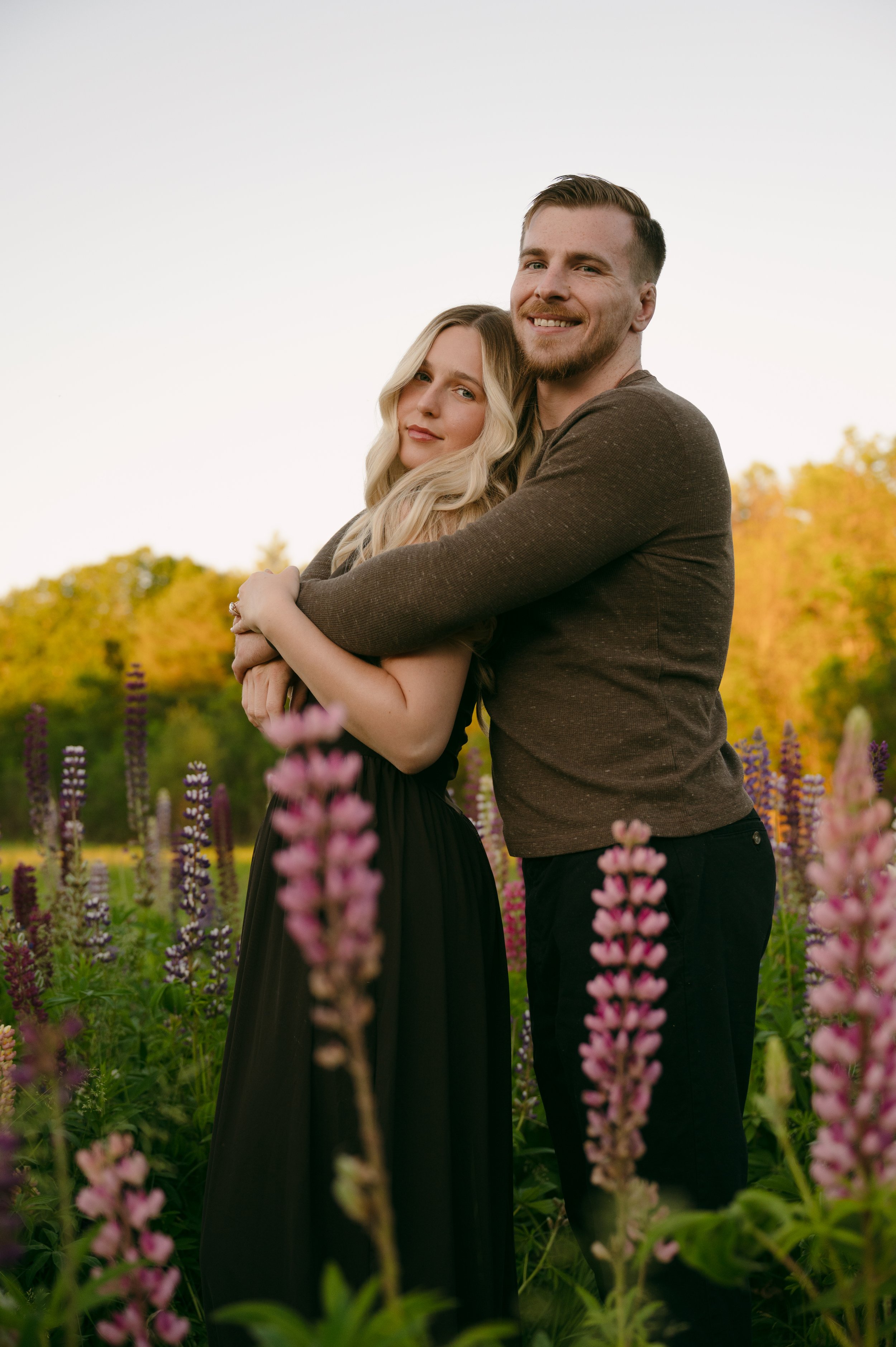 A young man and woman embracing in a garden with purple flowers and trees in the background, during sunset.