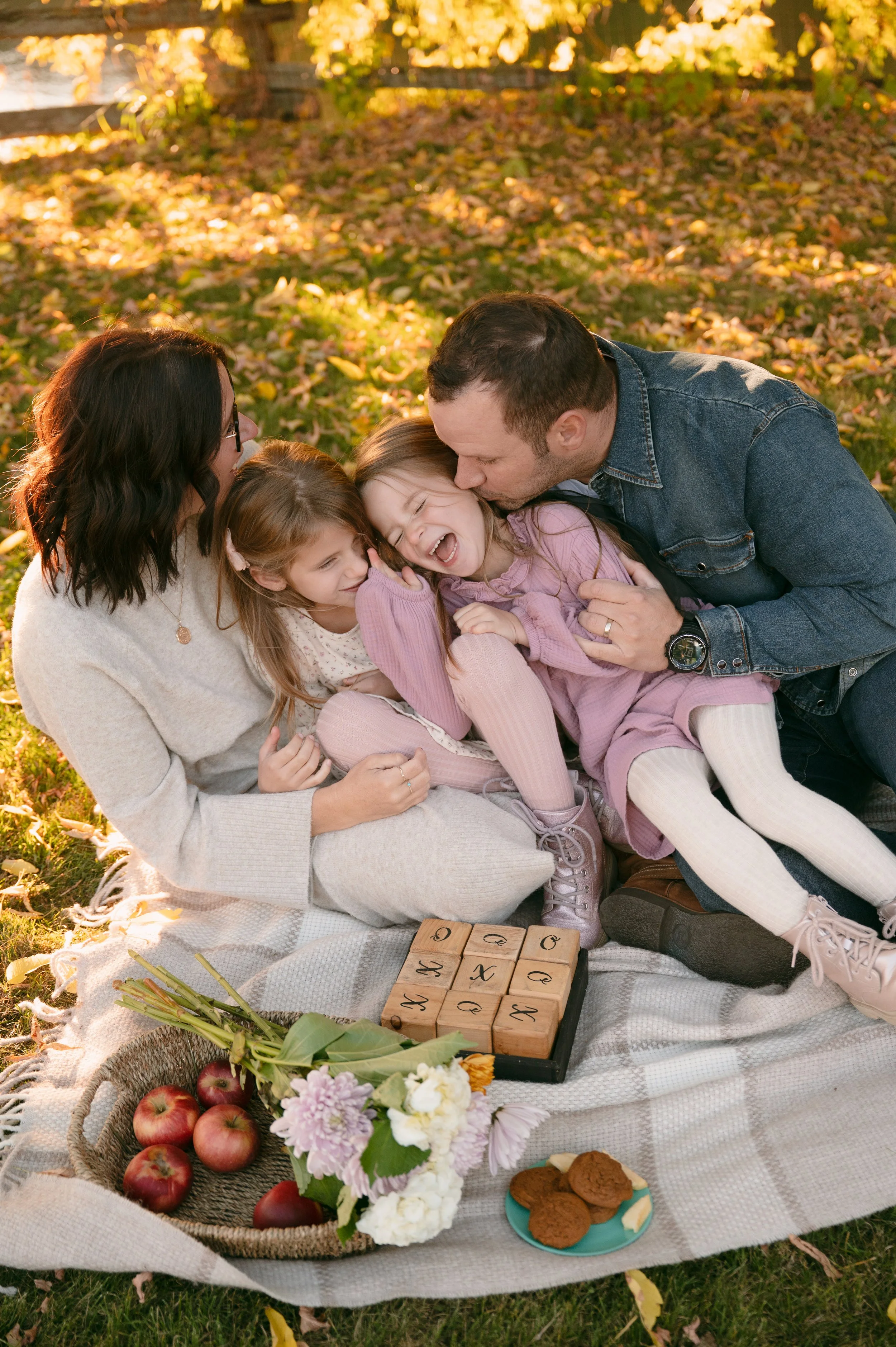 A family of five enjoying a picnic outdoors on a fall day, sitting on a blanket with apples, flowers, and cookies, with a backdrop of autumn leaves.