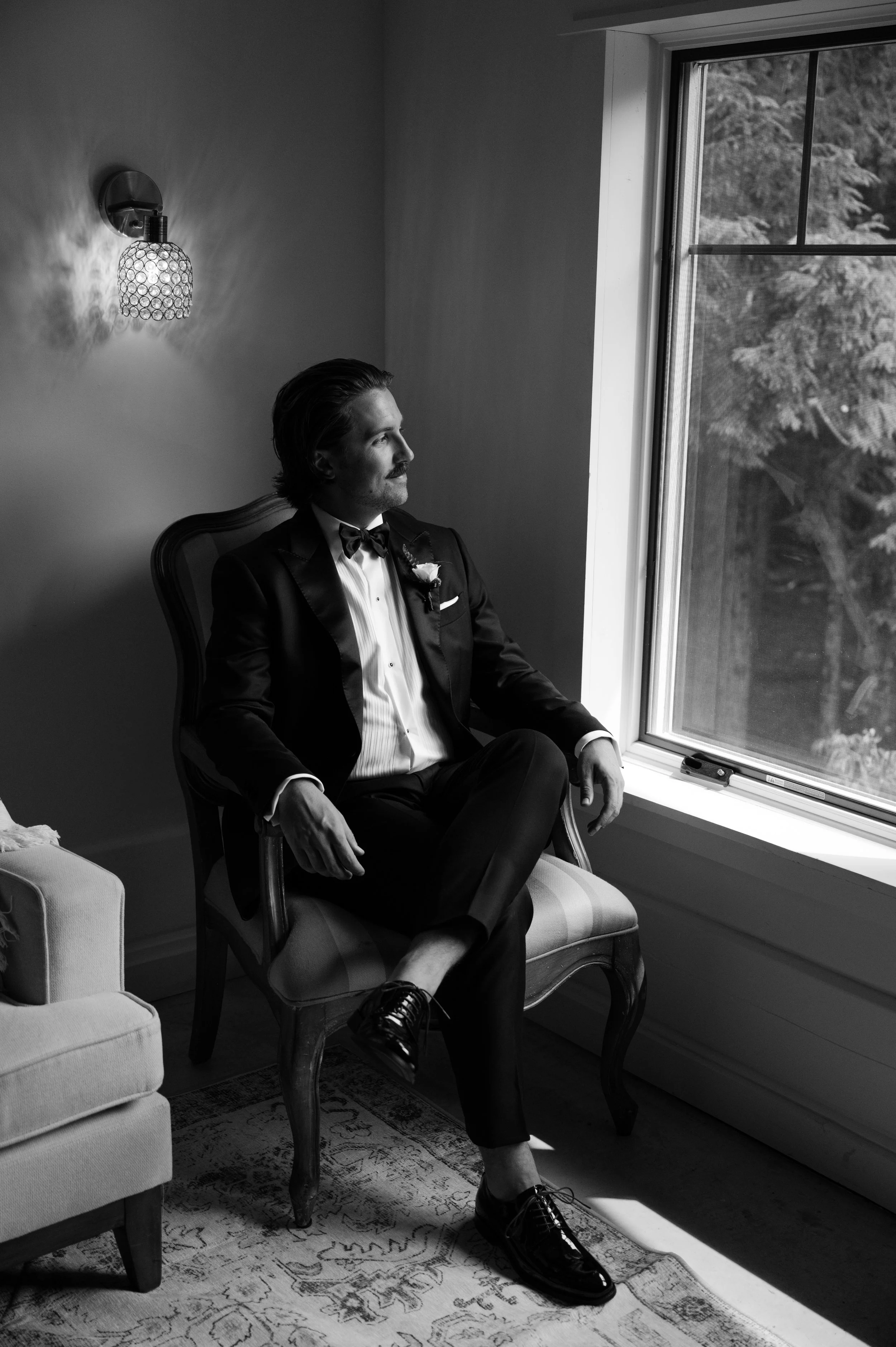 A man in a tuxedo sitting on a vintage wooden chair, looking out a window, with sunlight casting shadows in a room.