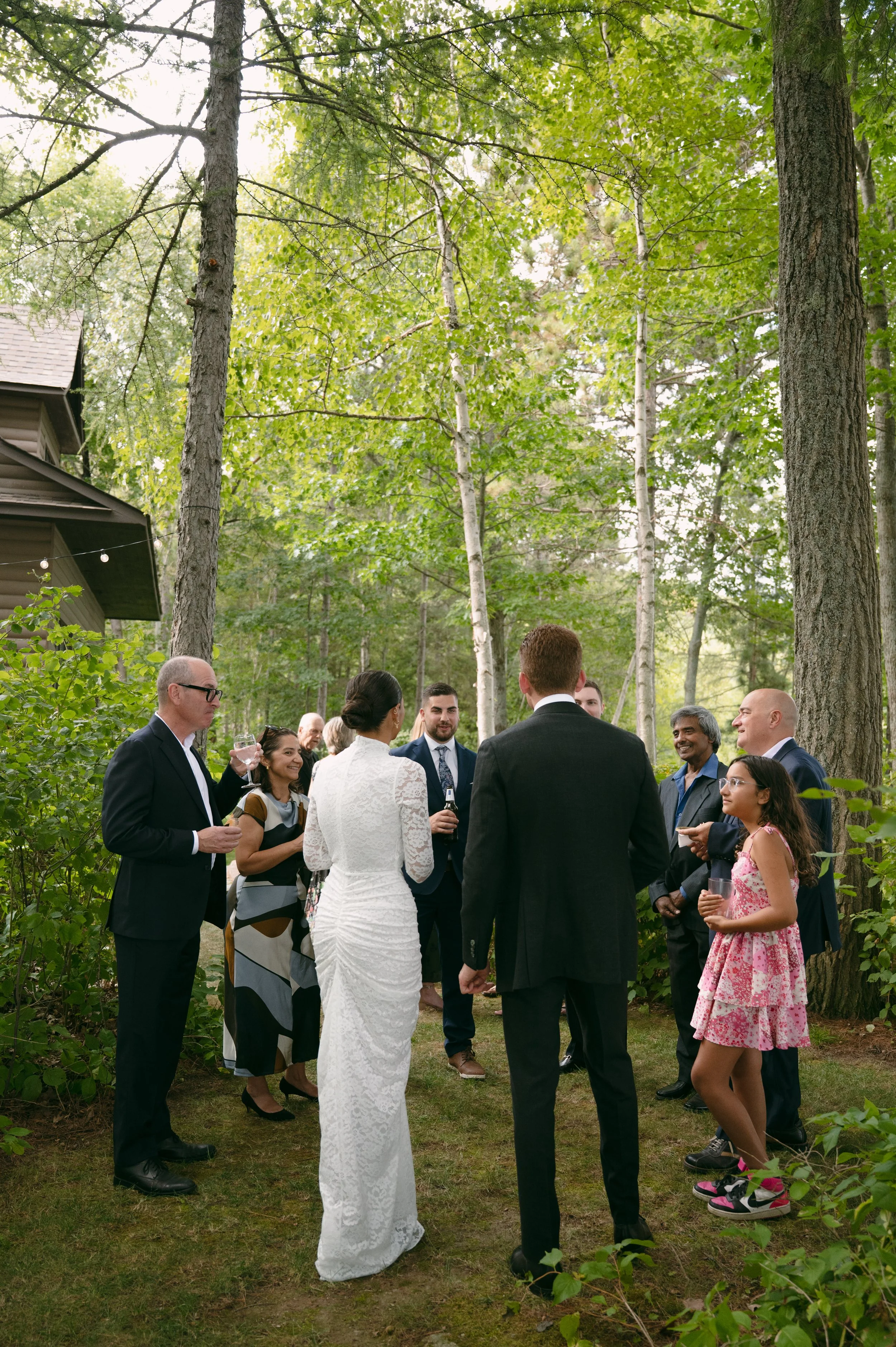 A group of people dressed in formal and semi-formal attire gathered outdoors among green trees, possibly at a celebration or reception.