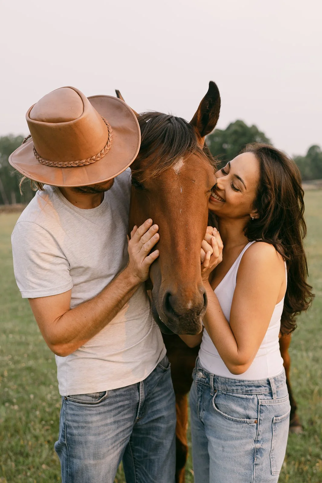 A man and woman hugging a brown horse outdoors in a grassy field, with trees in the background.