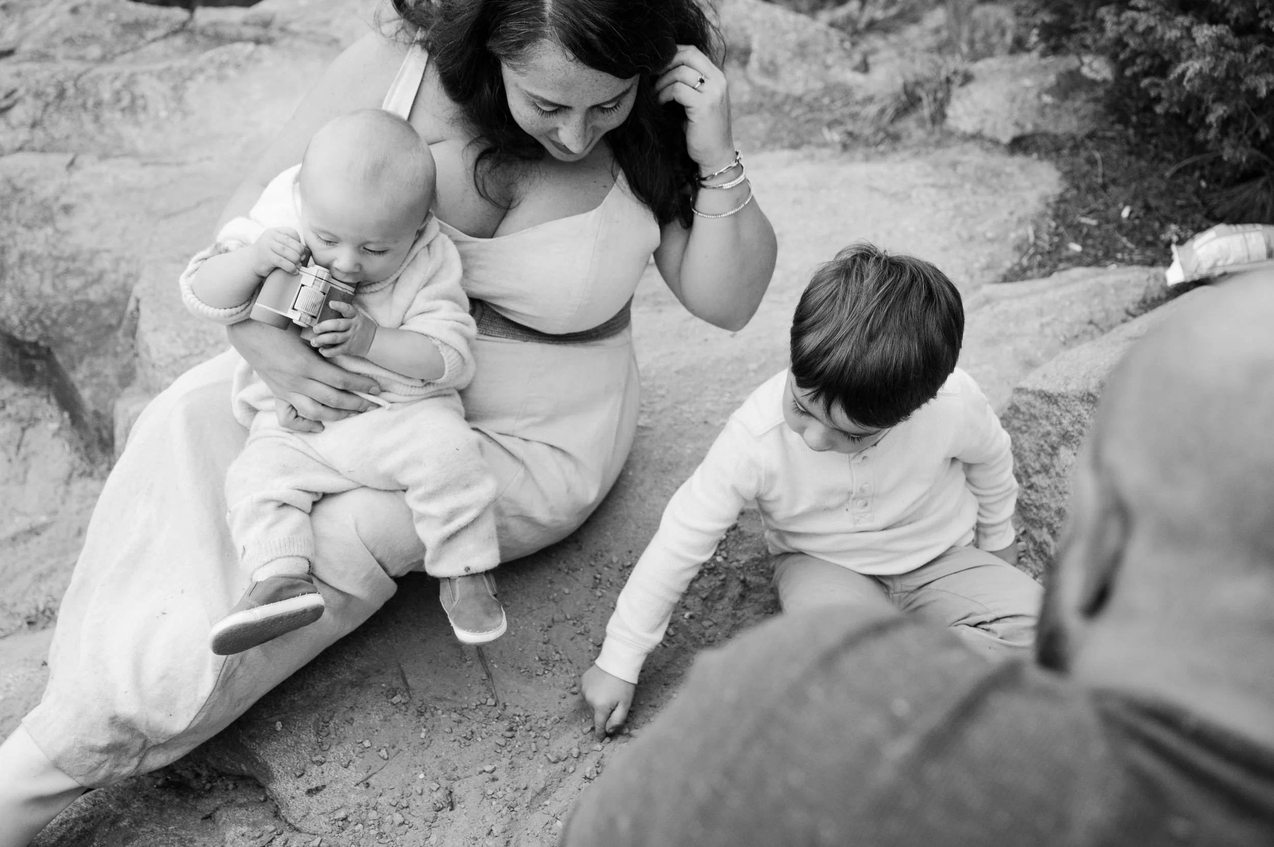 A woman with two children sitting outdoors on the ground. One child is a baby holding a camera, and the other is a young boy looking at the ground.