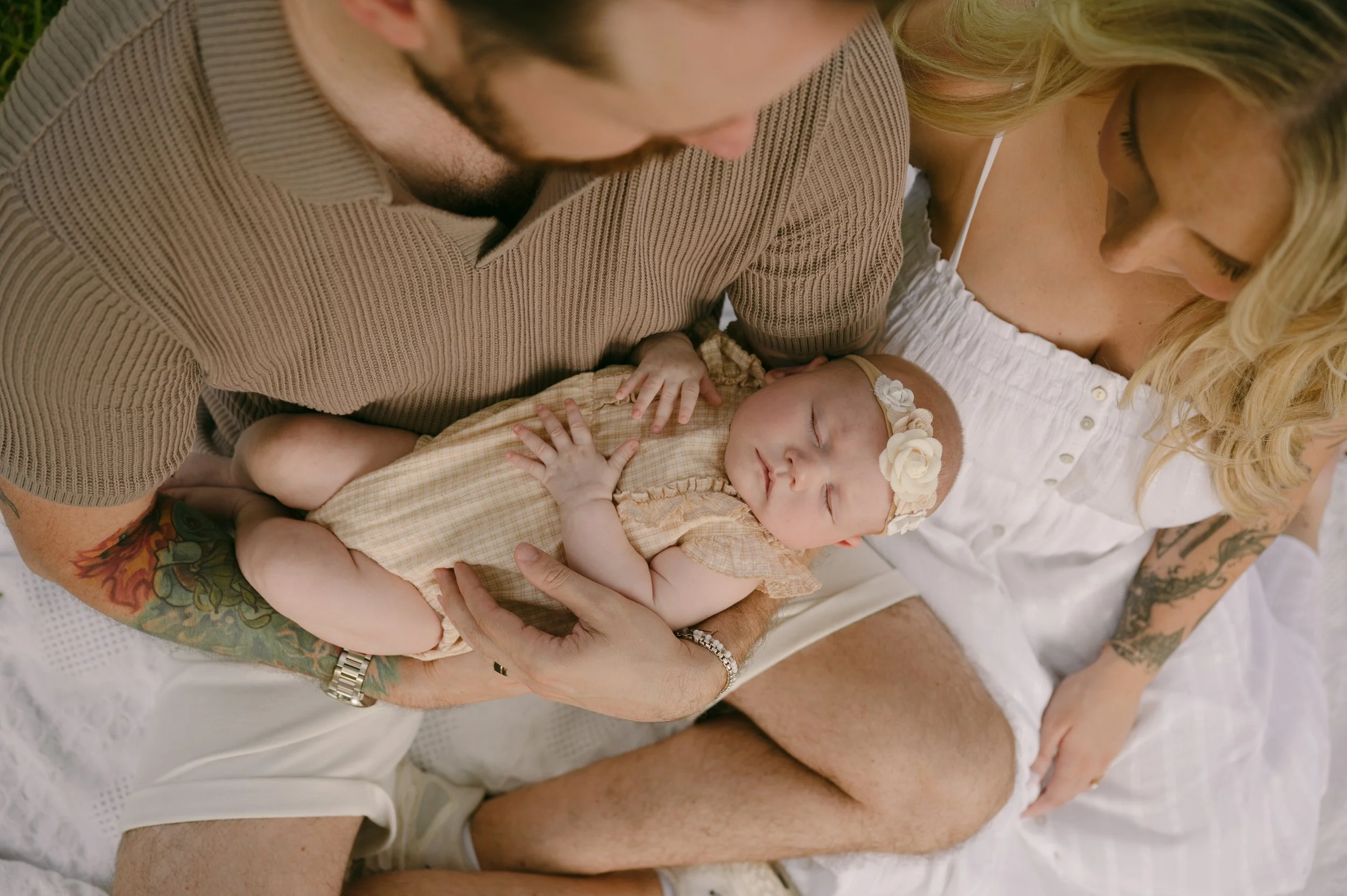 A man and woman are holding a sleeping baby girl on a bed, with the man in a brown shirt and the woman in white. The baby is wearing a beige dress and a floral headband.