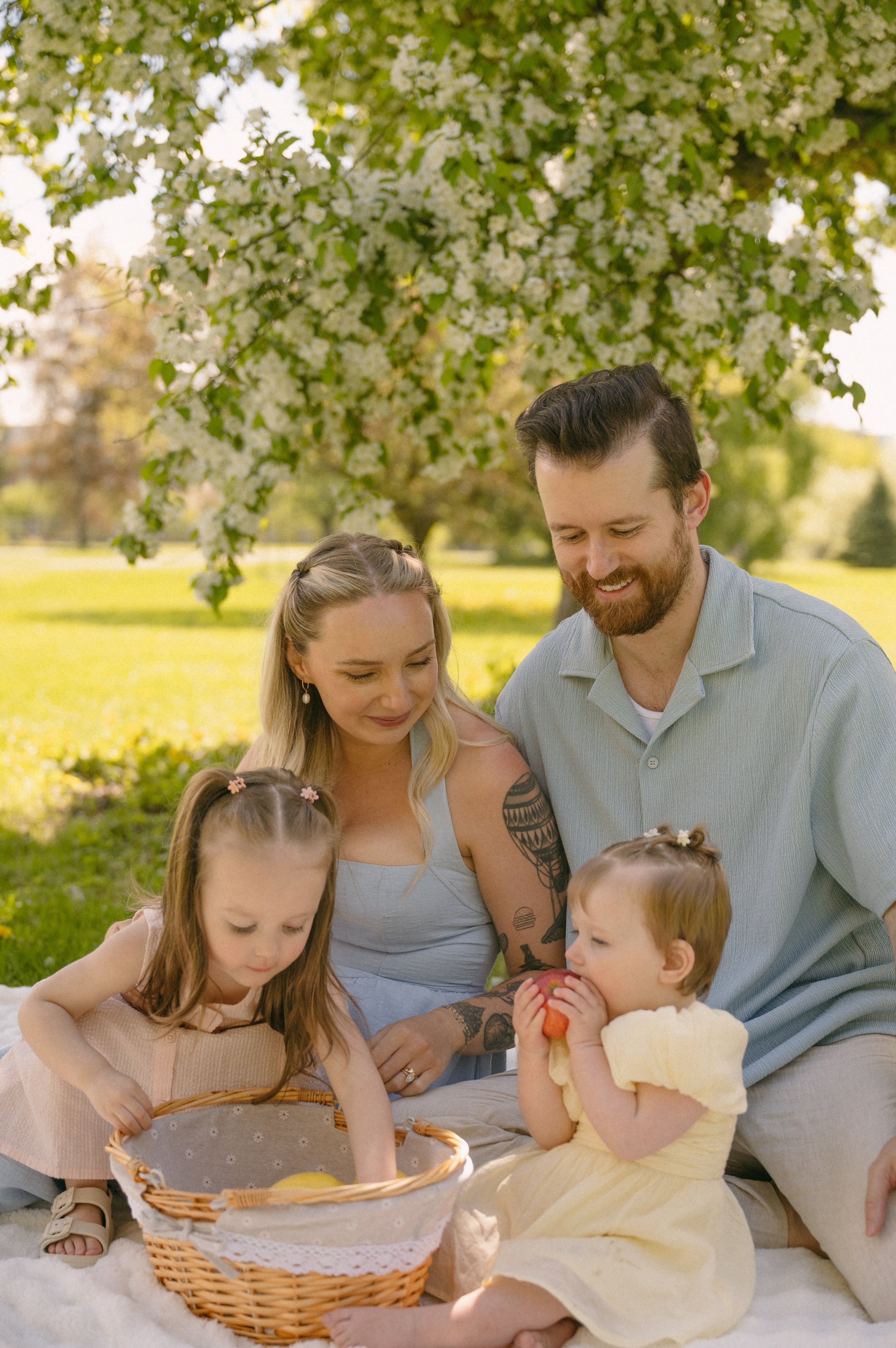 A family gathers outdoors under a flowering tree during spring, with children and adults engaging with a basket of apples.