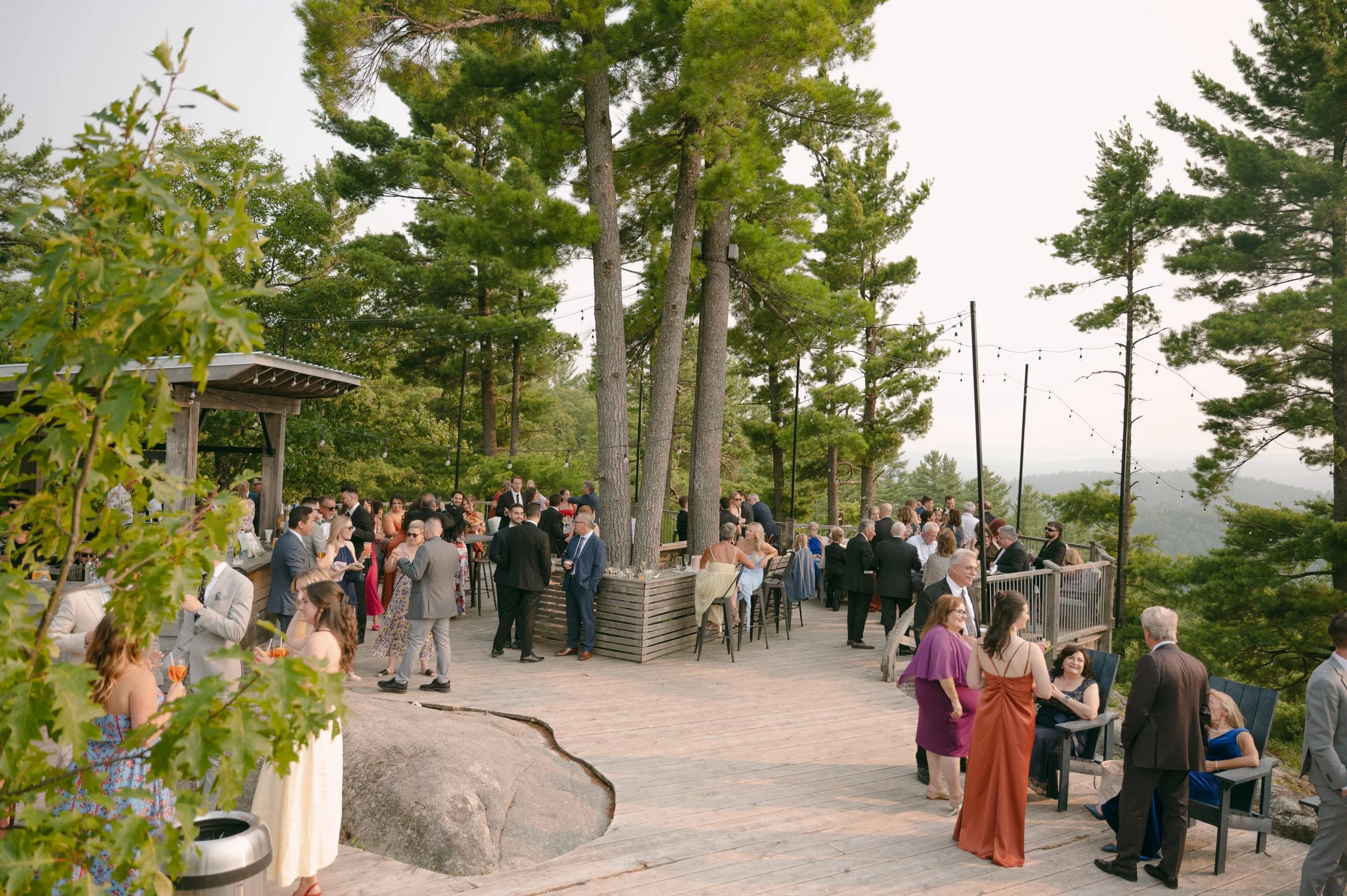 Guests socializing on a wooden outdoor terrace surrounded by tall trees during a gathering or celebration in the evening, with string lights overhead.