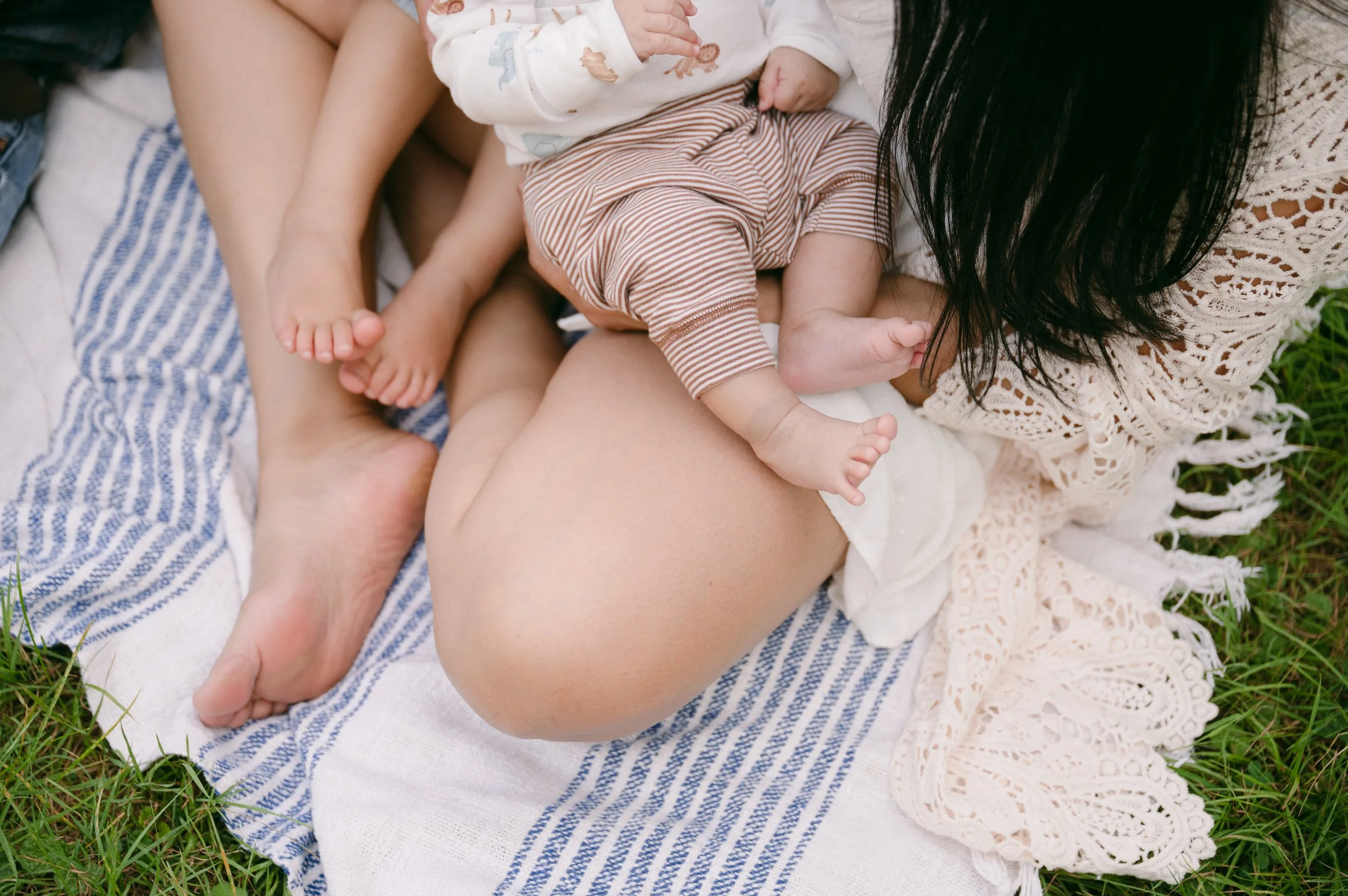 A woman and a baby lying on a blanket outdoors, with the woman breastfeeding the baby, surrounded by grass.