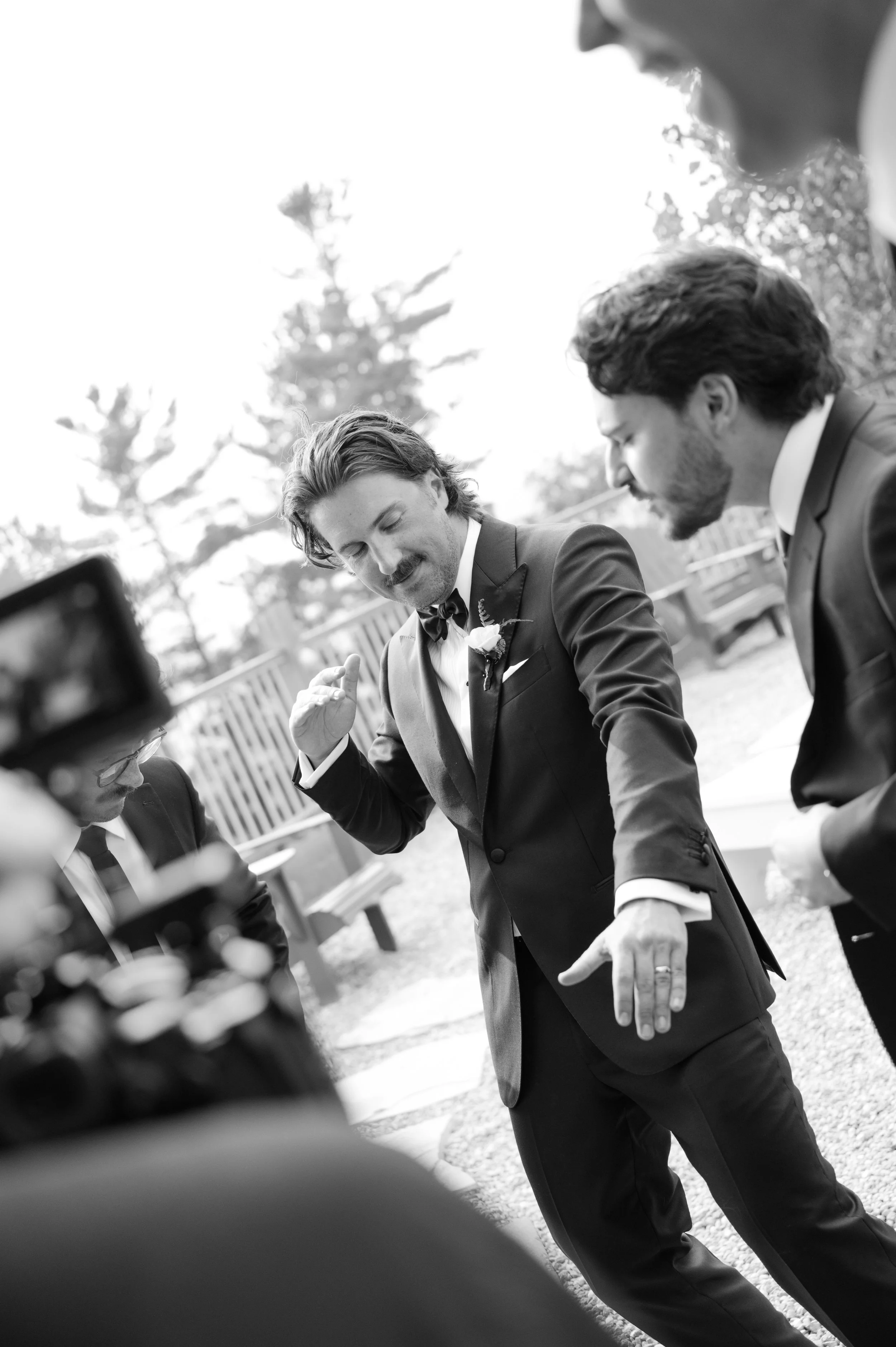 Black and white photo of a man in a tuxedo with a boutonniere, possibly at a wedding, speaking or gesturing with others around him.