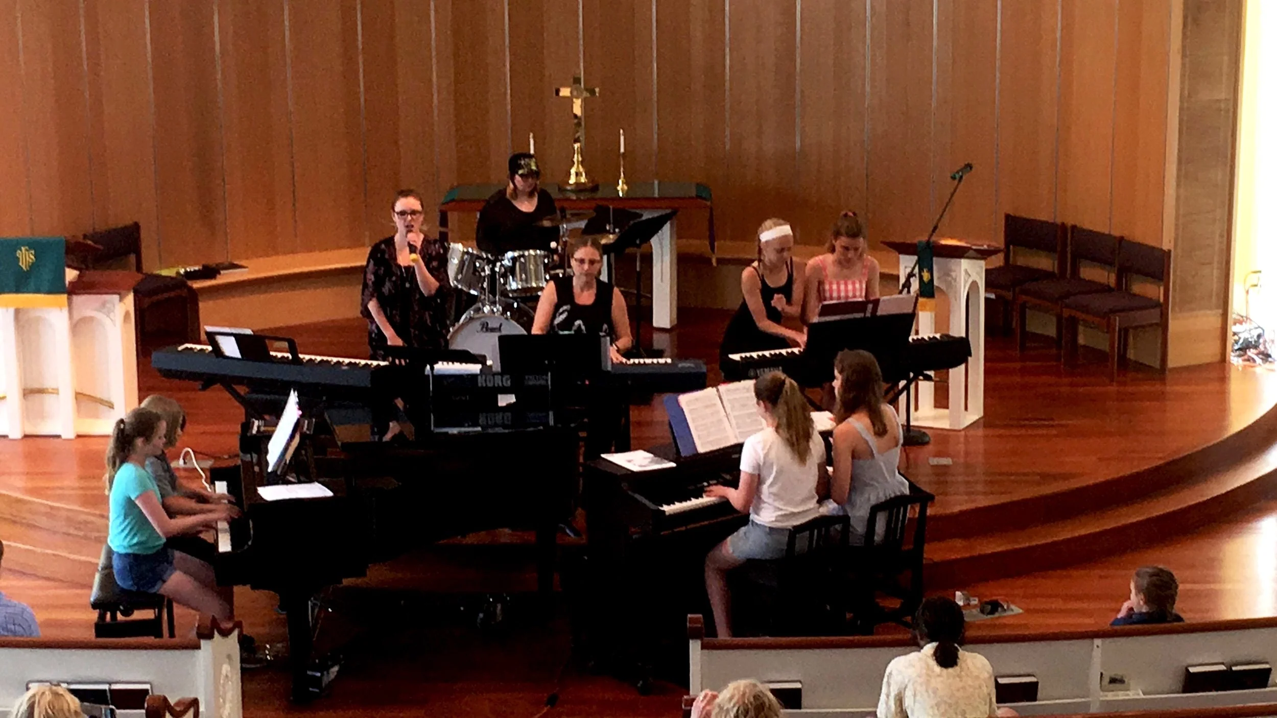 A group of young girls rehearsing in a church choir with musical instruments, including pianos and drums, on a wooden stage.