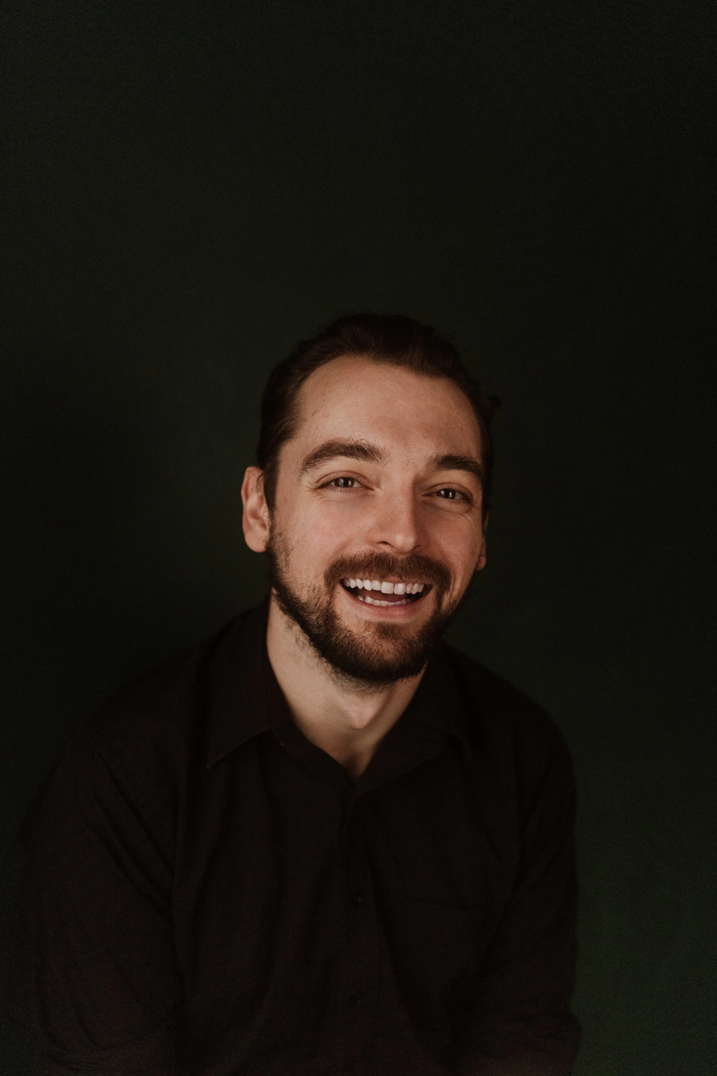 A smiling man with dark hair and a beard wearing a black shirt against a dark background.