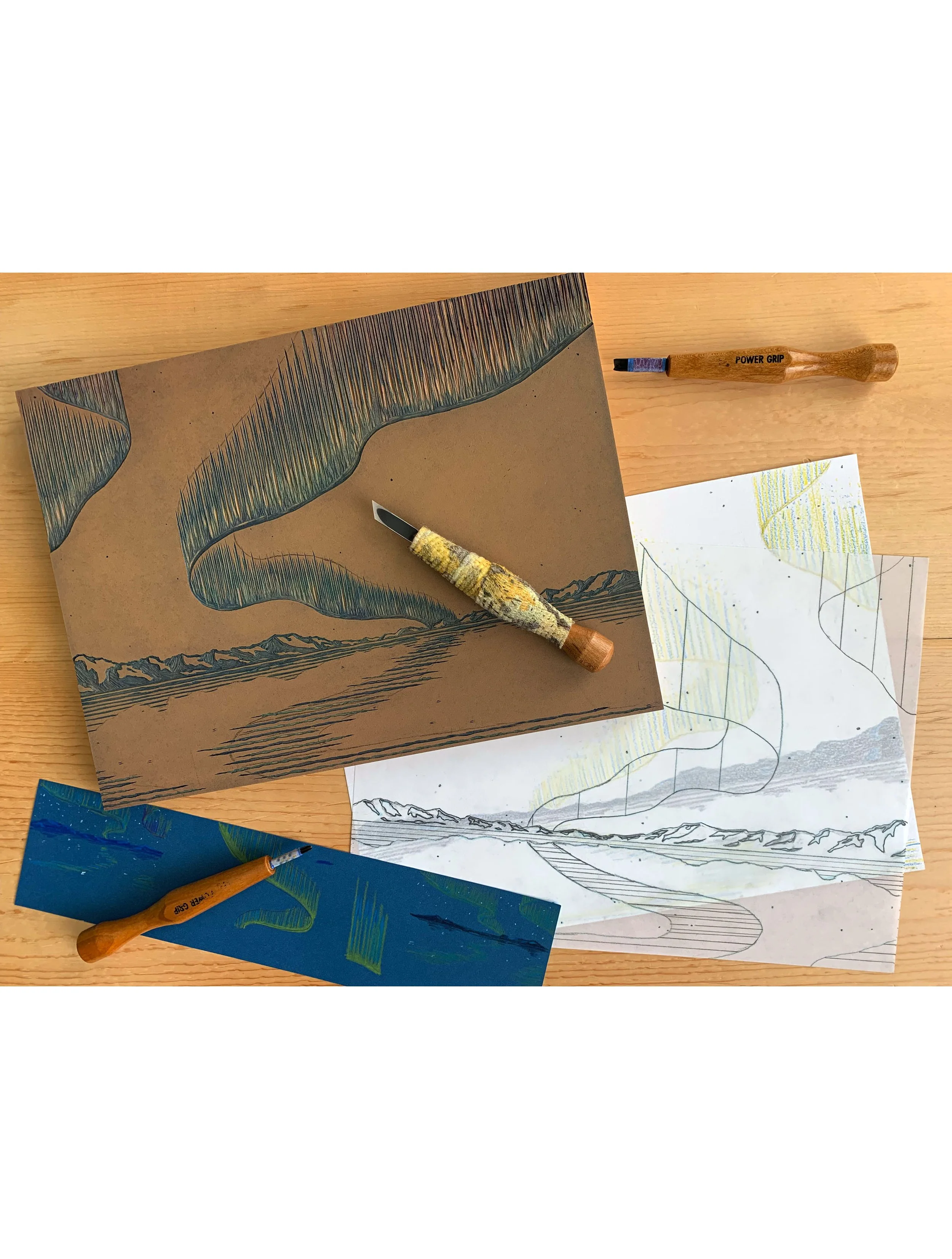 The carved lino block used to make the last layer of the handmade reductive linocut print of a northern lights landscape at night, in shades of blue. It lays amongst various sketches and tools used.