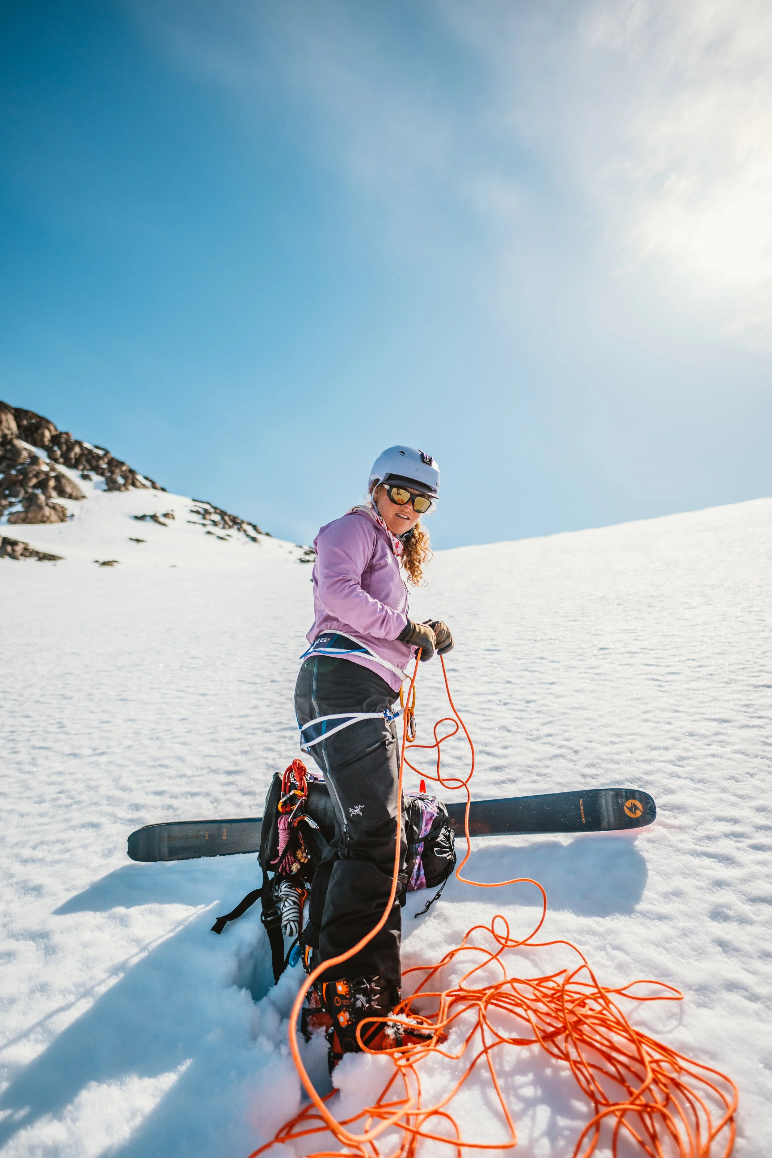 A woman in ski gear, including a helmet and sunglasses, standing in the snow with a snowboard and a tangled orange rope, outdoors on a snowy mountain slope.