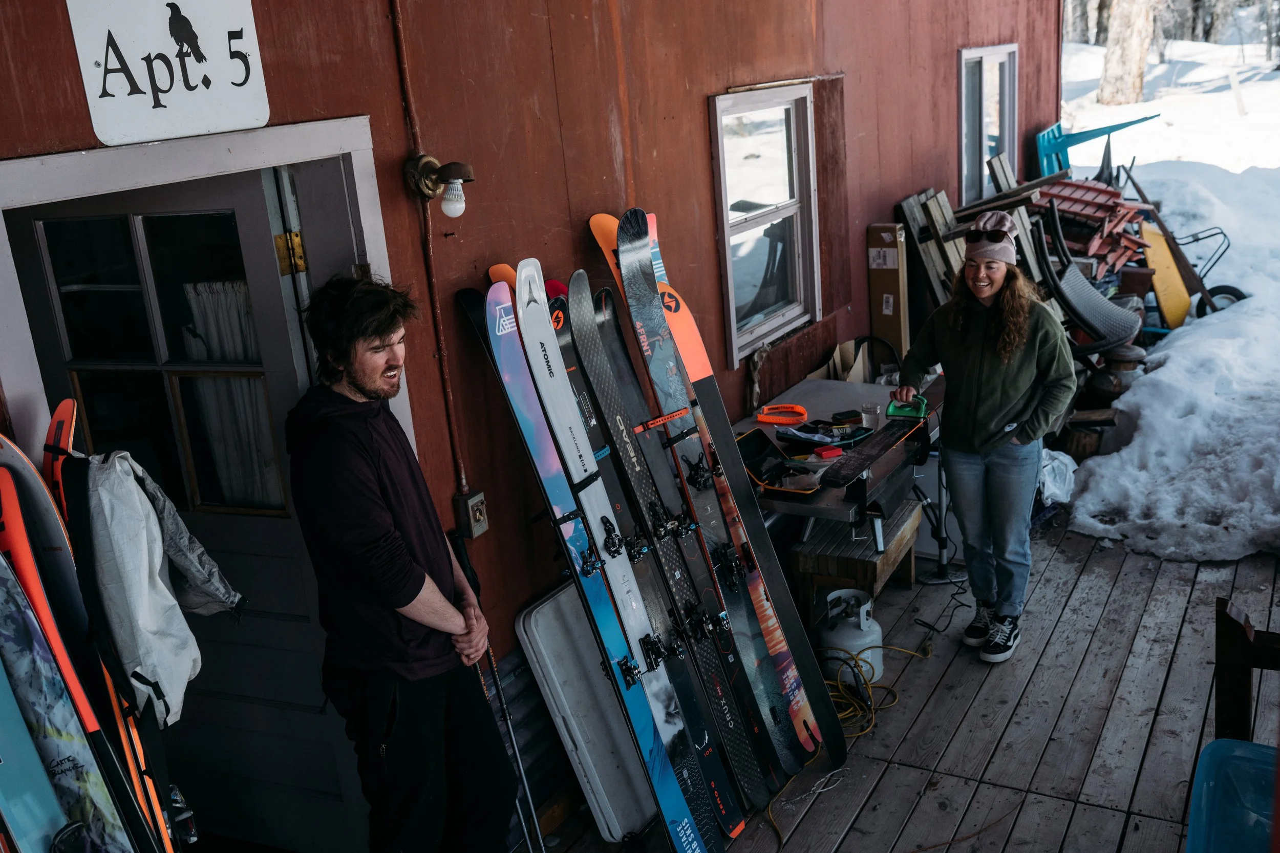 Two people preparing skis outside a wooden cabin in a snowy setting, with ski equipment and tools on a workbench.