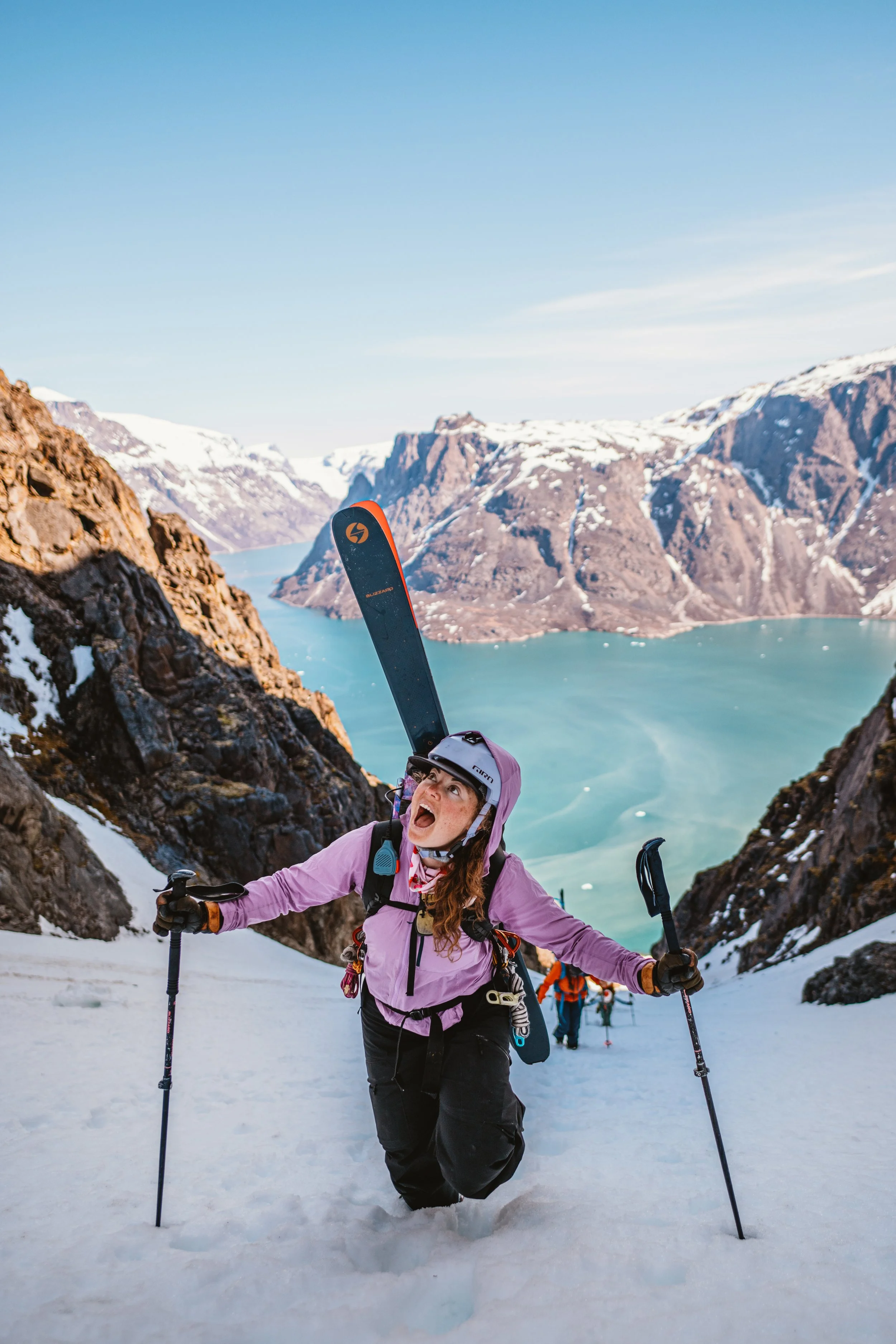 A woman hiking up snowy mountain terrain, carrying skis on her shoulder, with a lake and snow-capped mountains in the background.