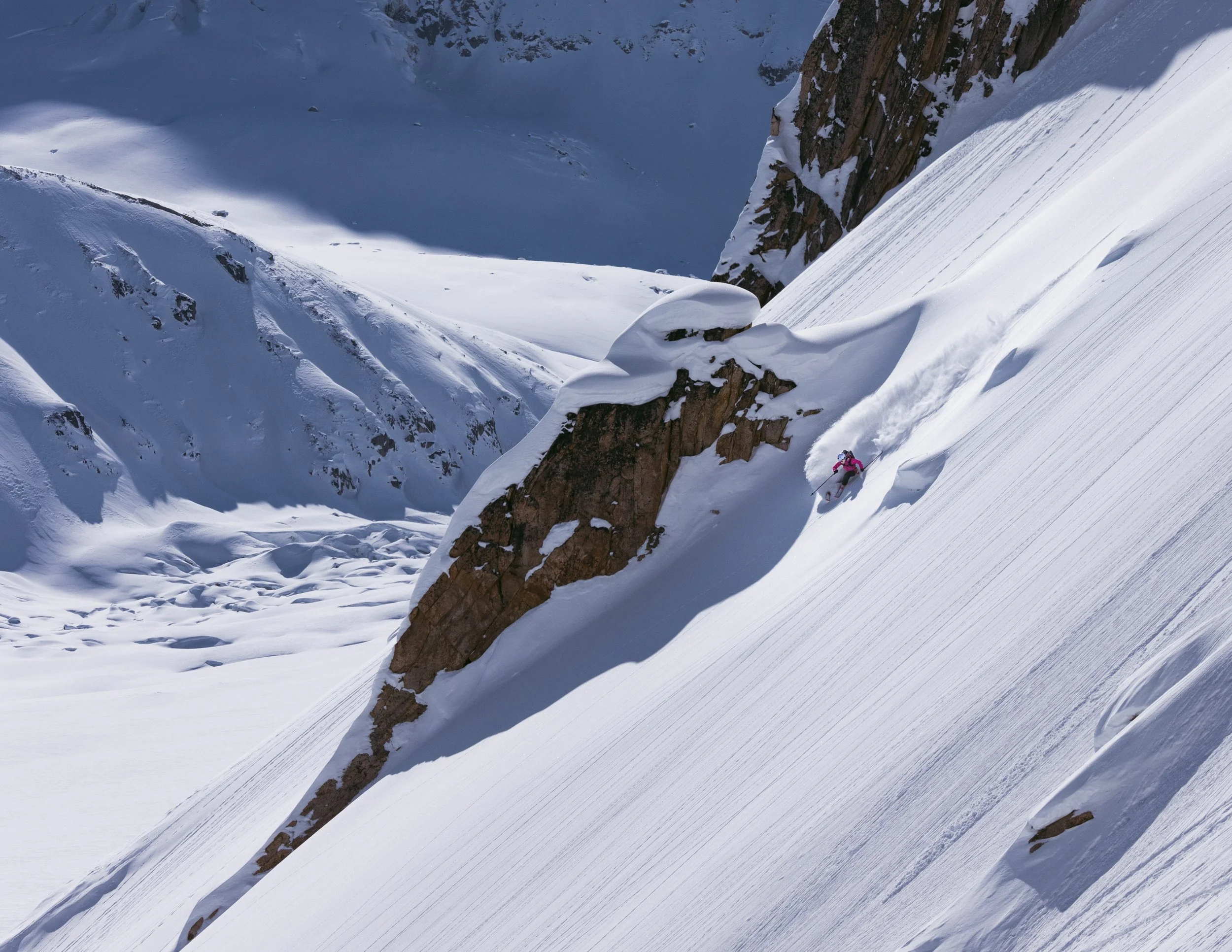 A skier in pink gear descending a steep, snow-covered mountain slope with rocky outcroppings and a valley in the distance.