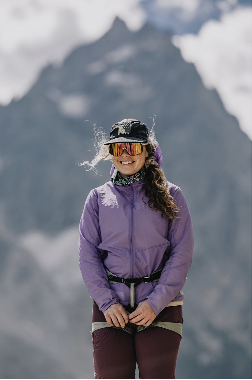 A woman in outdoor hiking gear, wearing a purple jacket, maroon pants, and reflective sunglasses, standing in front of a mountainous landscape with clouds.