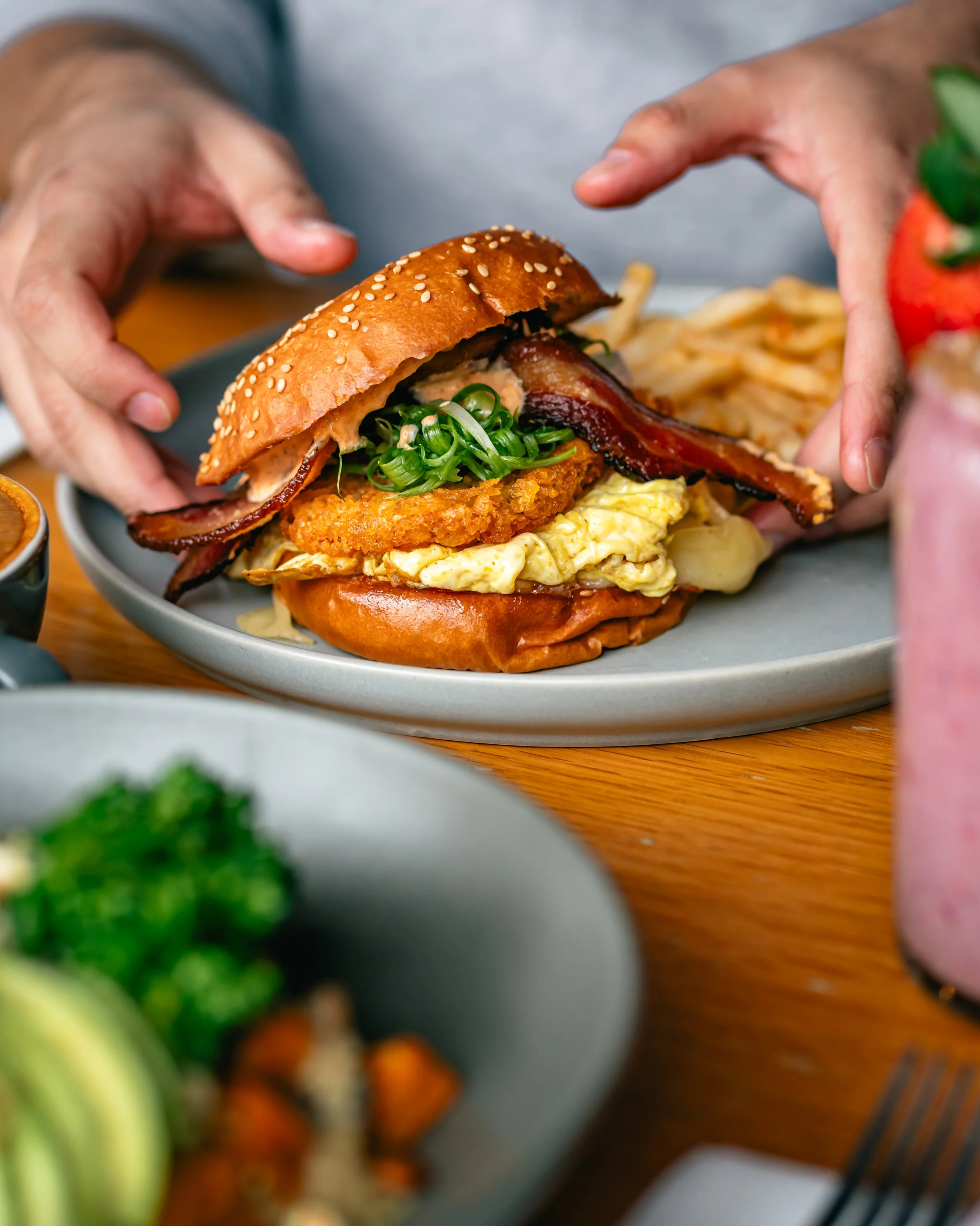 Person holding a burger with bacon, fried chicken, scrambled eggs, green onions, and a sesame seed bun, with fries on a plate, surrounded by sides and drinks.