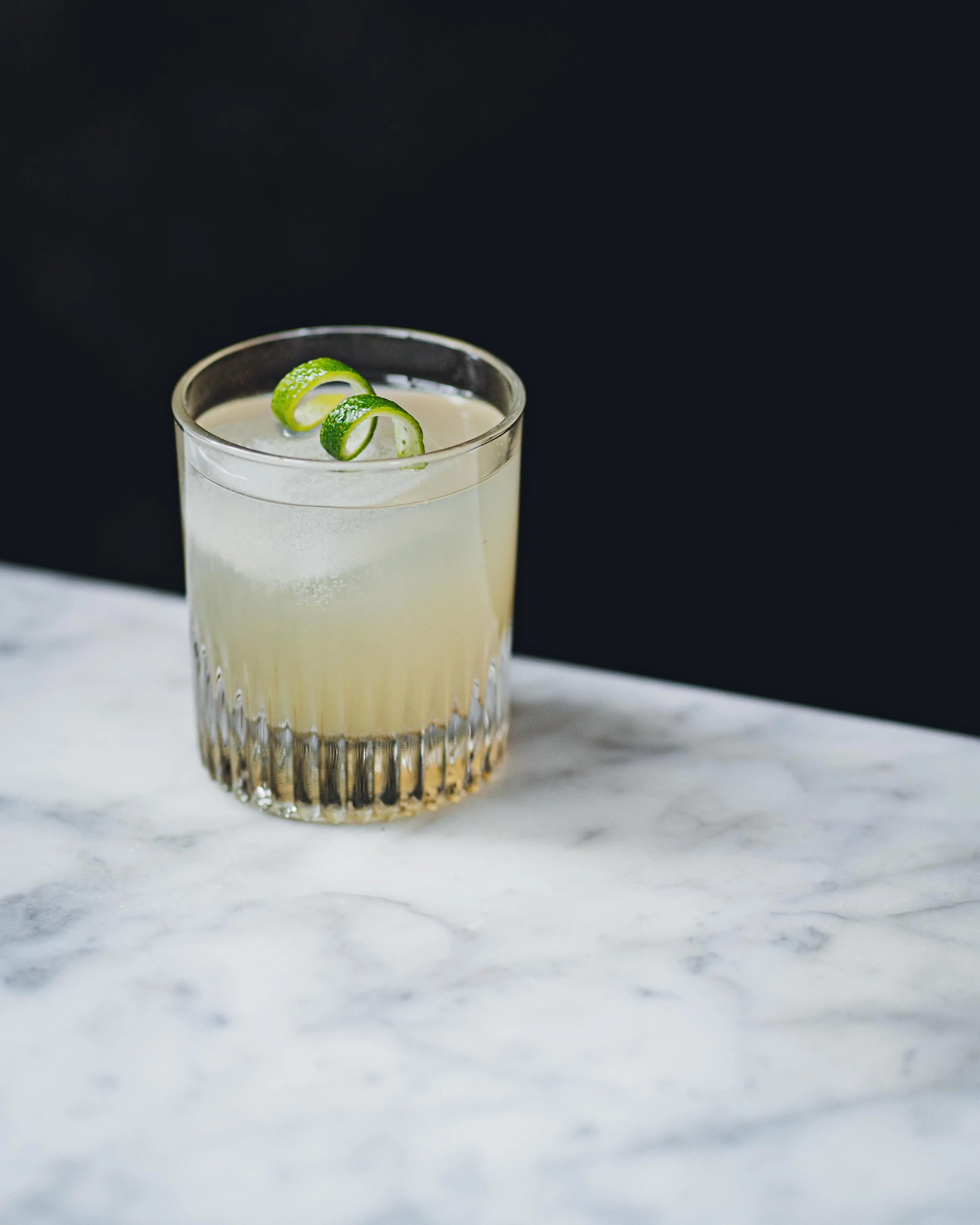 A glass of pale yellow cocktail garnished with a twisted lime peel, placed on a white marble surface against a dark background.