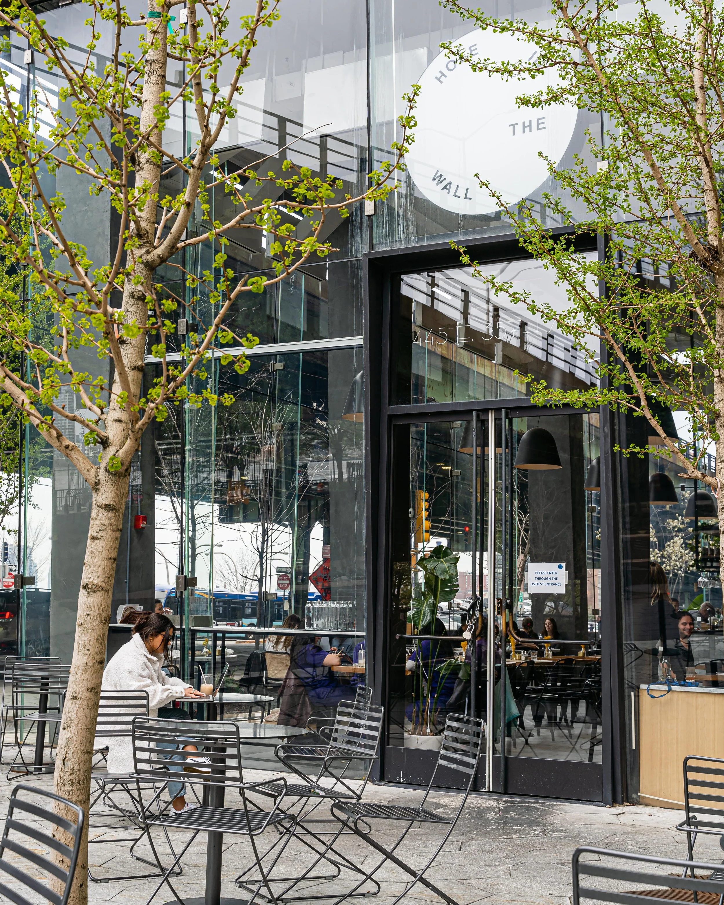 A modern glass-front cafe with outdoor seating, some patrons working or socializing inside, and trees with budding green leaves outside.