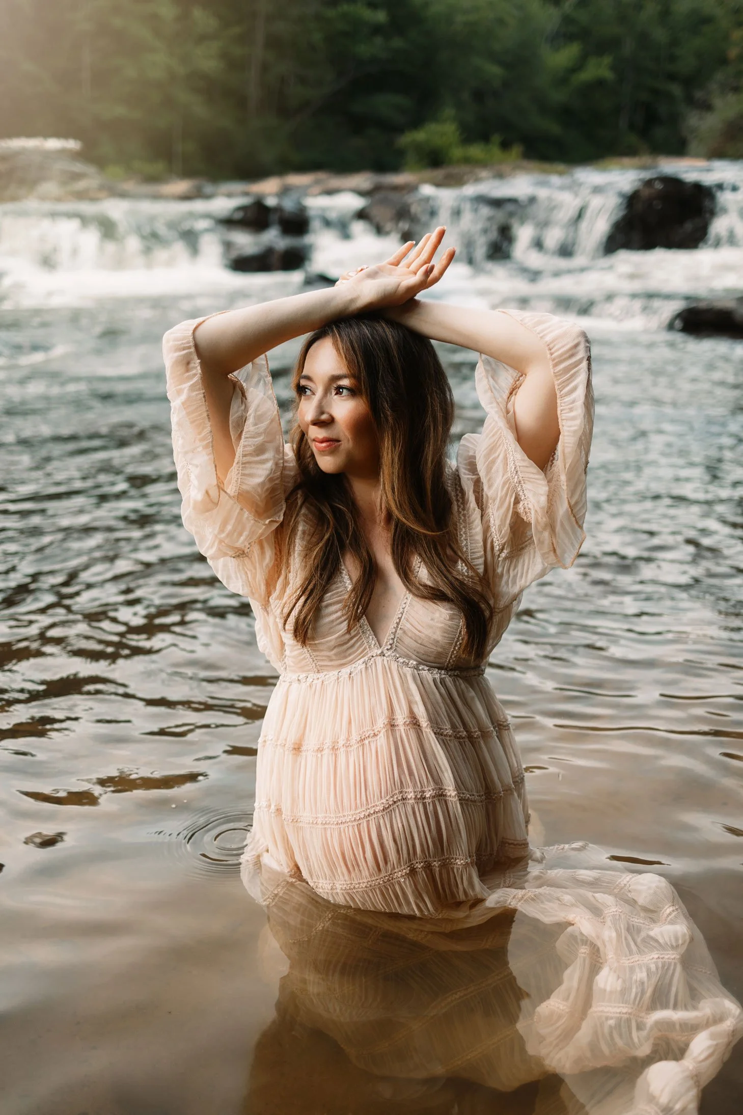 Pregnant woman standing in a North Georgia creek with arms raised, wearing a boho maternity gown during a Dahlonega waterfall maternity session.