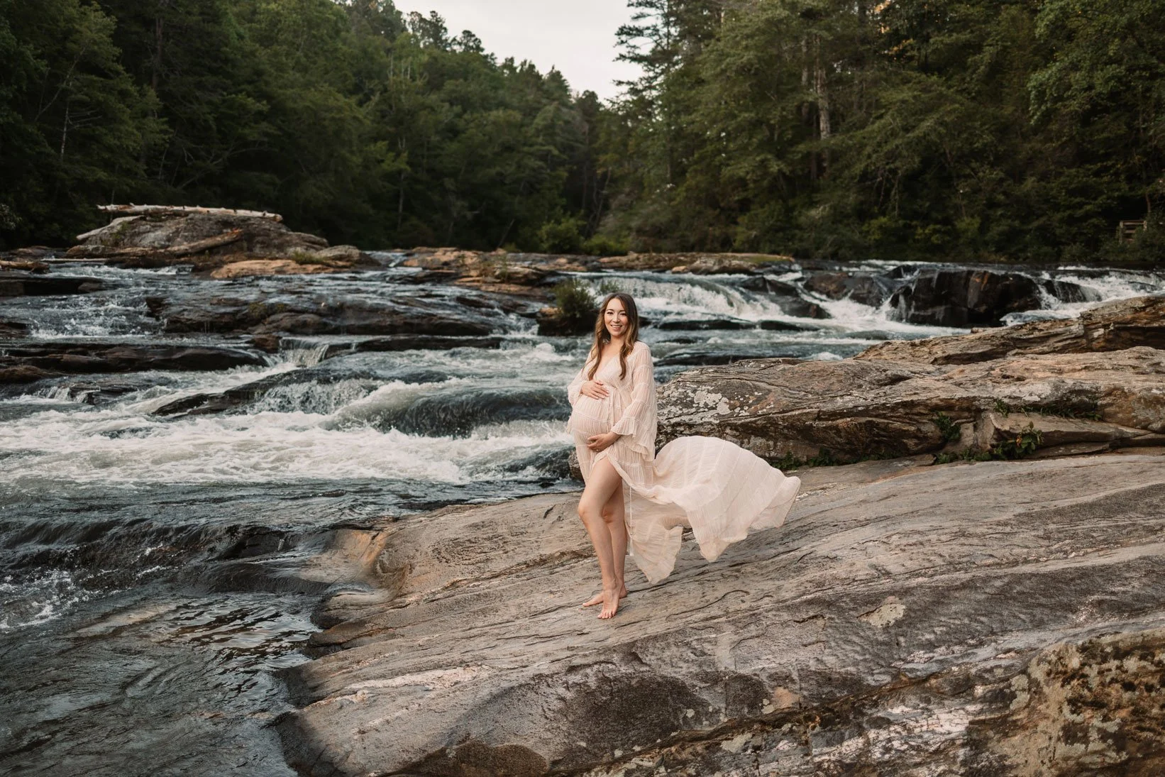 Expecting mother walking barefoot across river rocks beside a rushing creek near Dahlonega, Georgia during her outdoor maternity photoshoot.