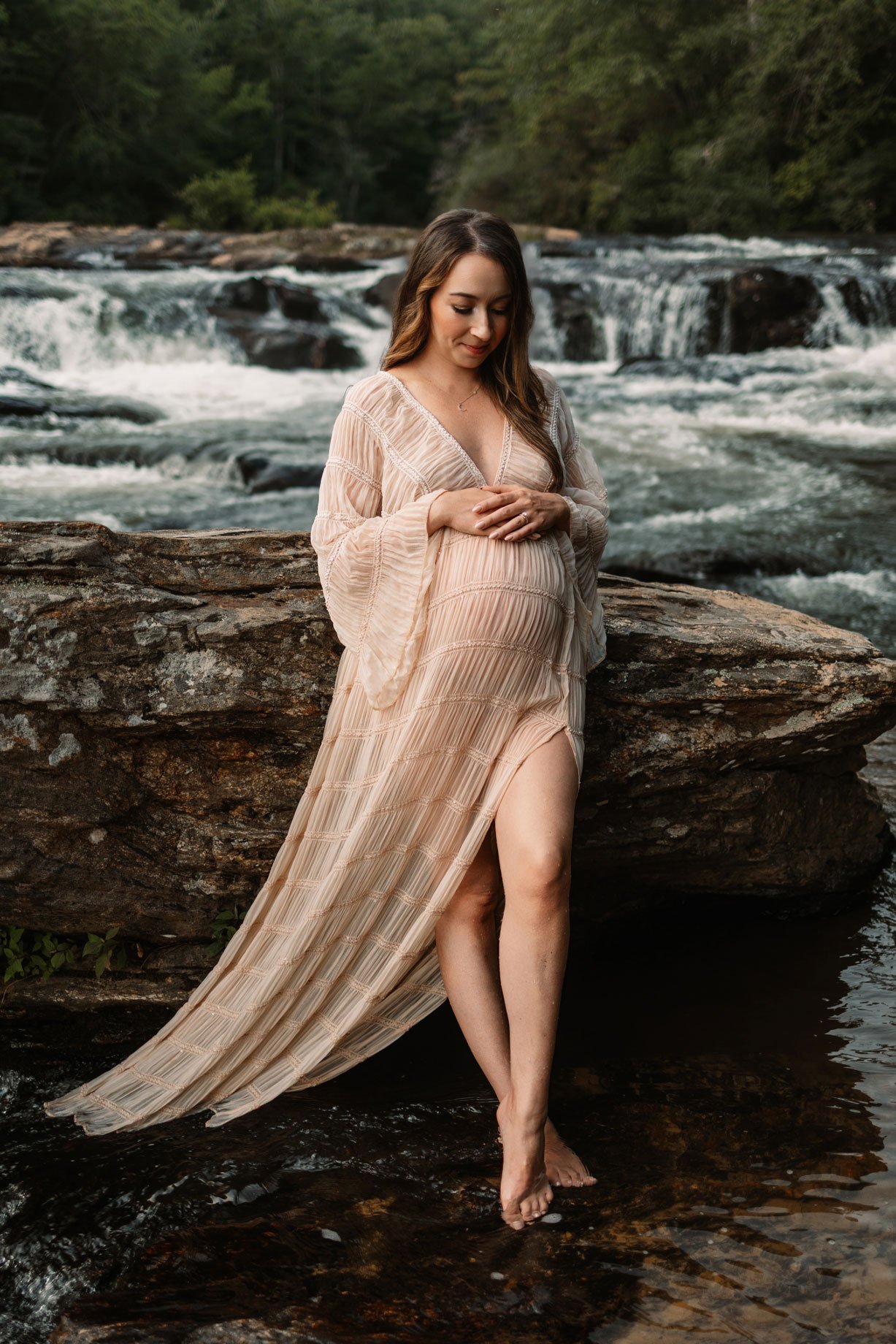 Boho maternity portrait of an expecting mother standing on rocks with a Dahlonega waterfall behind her, holding her belly during a North Georgia outdoor photoshoot.