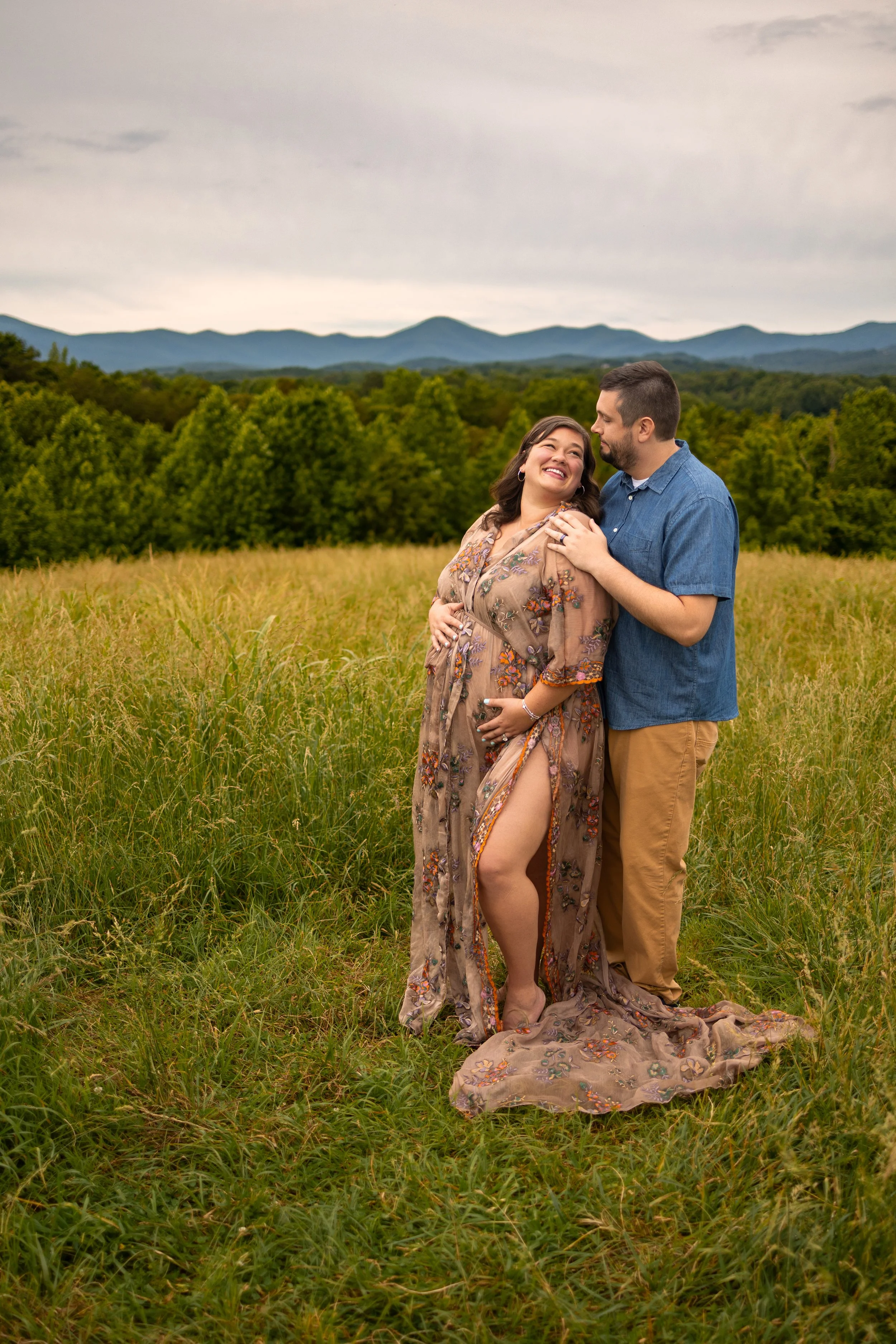 Expecting couple embracing in a grassy North Georgia field during a maternity photo session with mountain views in the background