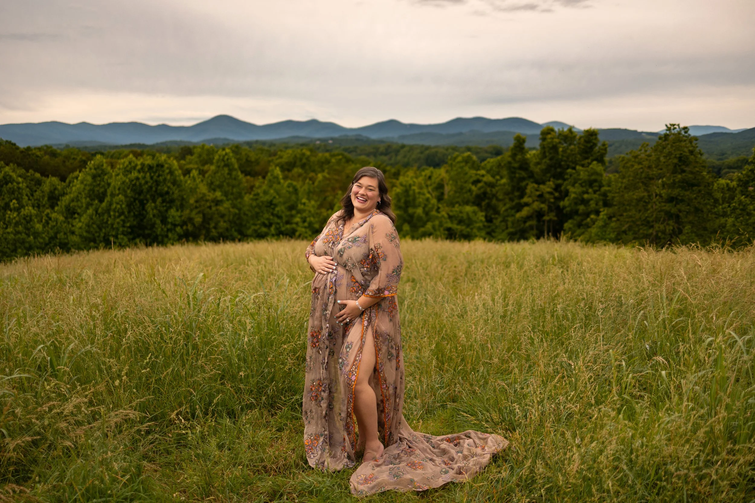 Joyful pregnant woman in a sheer floral gown standing barefoot in a grassy field with the Blue Ridge Mountains in the background. Buford Maternity photographers