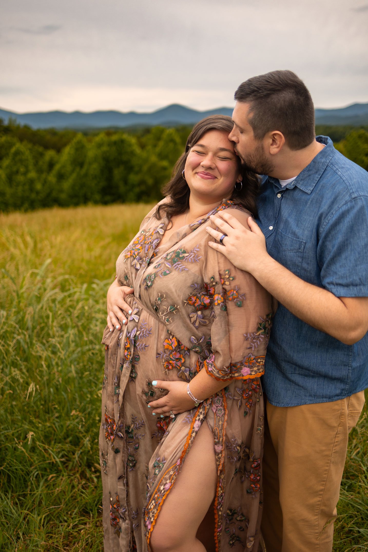Expecting couple embracing in a grassy North Georgia field during a maternity photo session with mountain views in the background