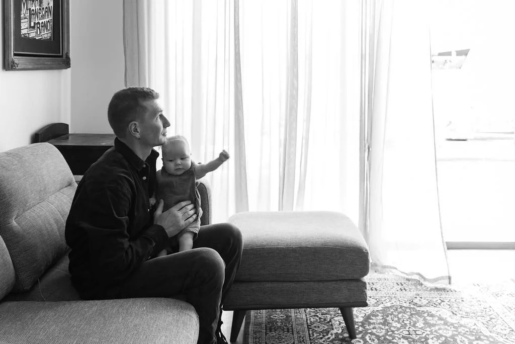A man sitting on a sofa holding a baby, looking out the window with sheer curtains, in a living room.