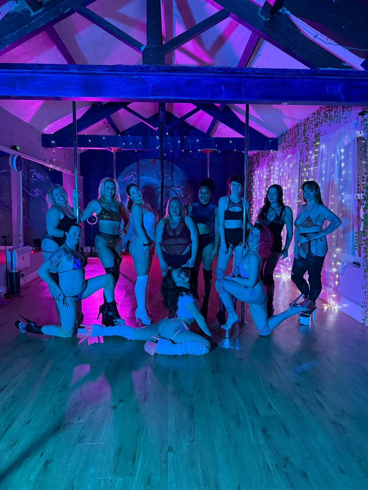 Group of women in lingerie and fishnets posing and kneeling on the wooden floor of a dance studio with purple and pink lighting and decorative string lights.